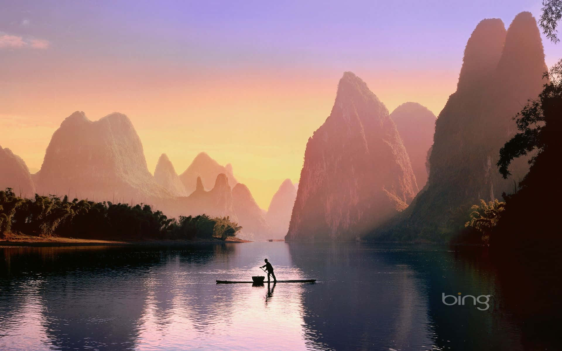 漁夫在平靜的河上划竹筏，夕陽映照山峰倒影 — 必應 壁紙