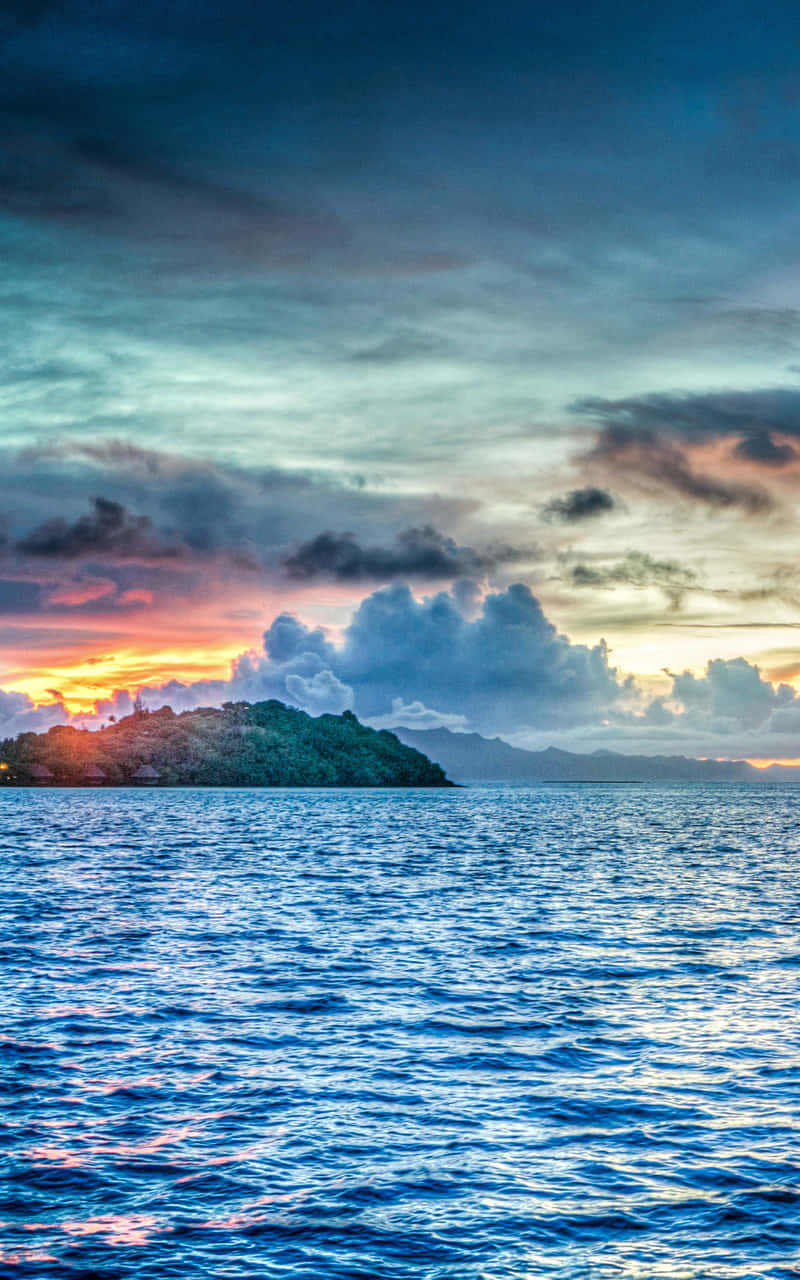 Bora Bora 桌布 壁紙 — 夕陽西下，天空被染成火橙色和深藍色，平靜的海洋上浮現出一個鬱鬱蔥蔥的綠色島嶼輪廓。