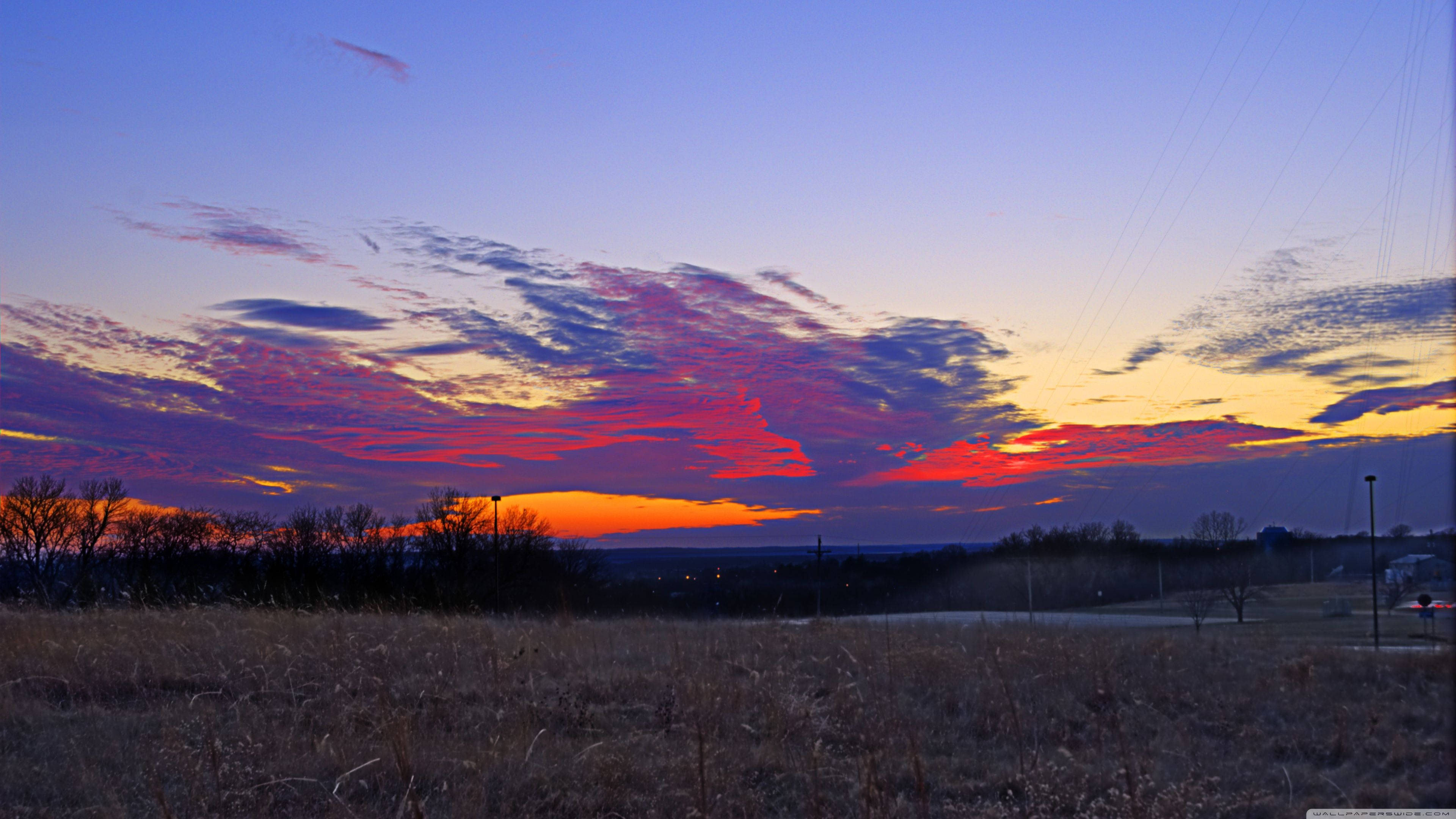 Kansas 桌布 壁紙 — 夕陽西下，天空被染成火紅、橙黃和紫色，映襯著乾草和遠樹的鄉村剪影。