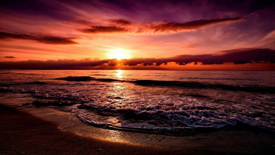 Beach Sunset HD 桌布 壁紙 — 夕陽西下，海面上火橙色和紫色的雲彩映照在濕沙和輕柔的海浪上，場面壯觀。