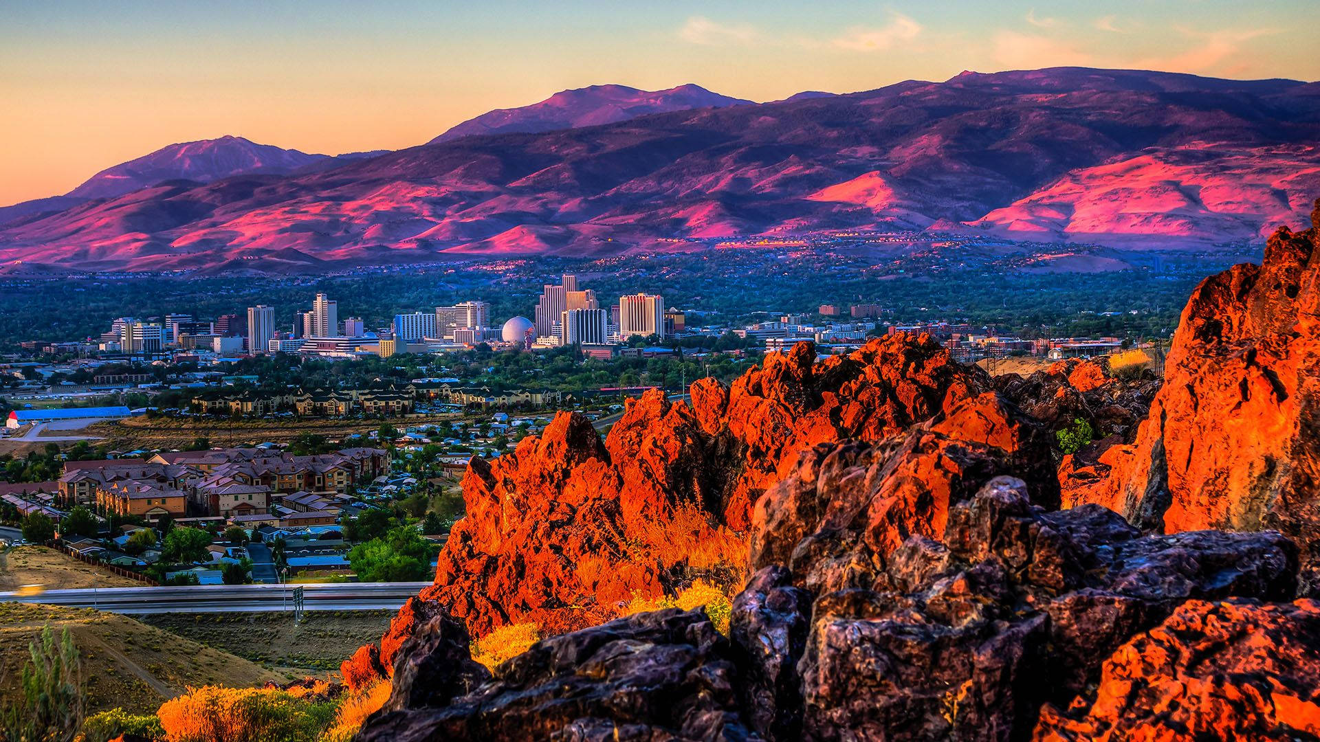 Photography Desktop 桌布 壁紙 — 夕陽西下，溫暖的色調灑在崎嶇的紅色岩石和遠山之上，城市在下方延伸。