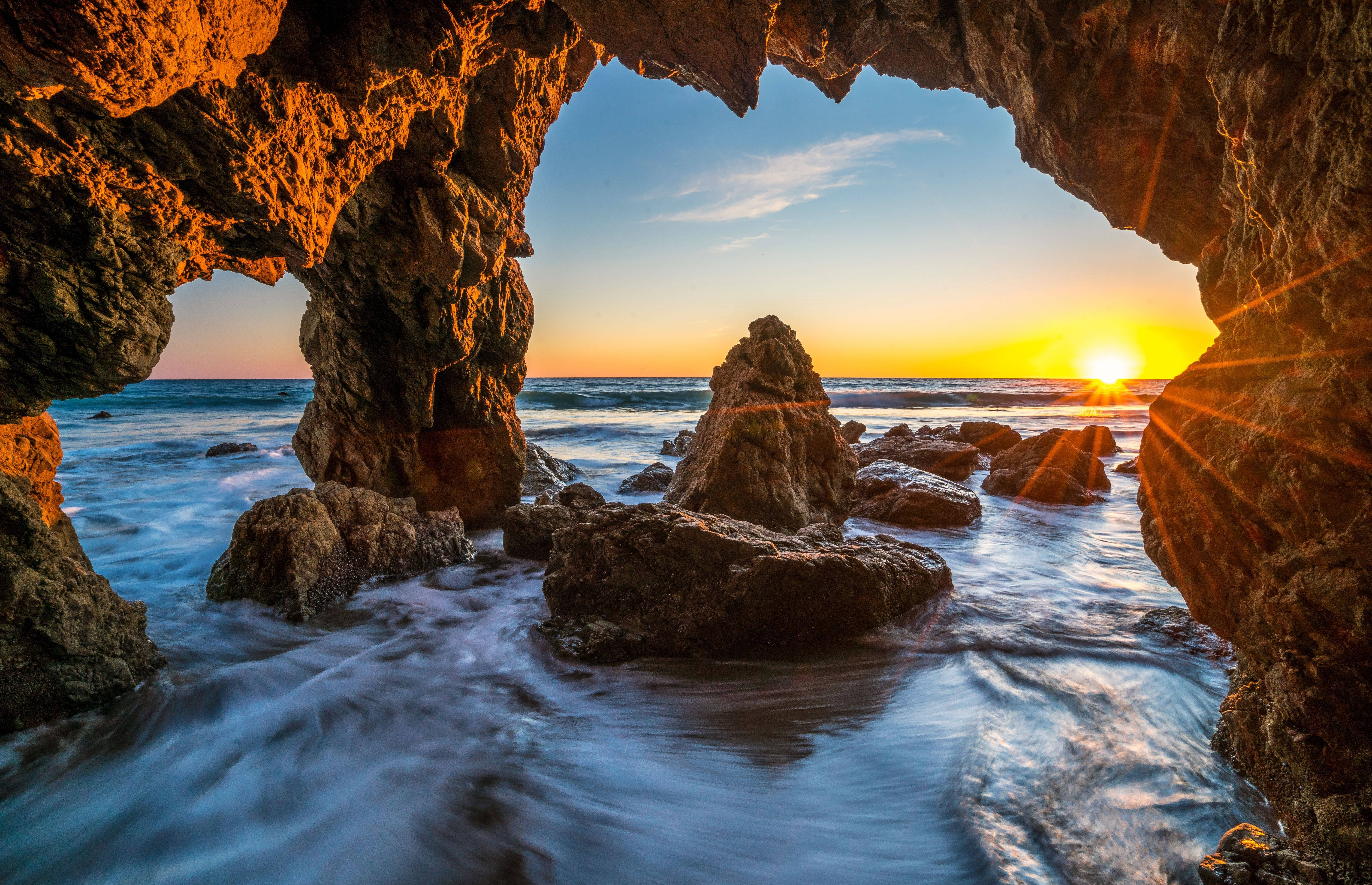 Malibu 桌布 壁紙 — 夕陽透過天然岩石拱門閃耀，框住拍打的海浪和崎嶇的海岸巨石。