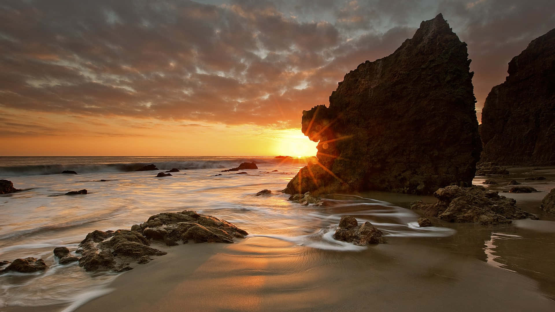 1920x1080 Malibu 桌布 壁紙 — 夕陽透過岩石拱門閃耀，海浪在戲劇性的多雲天空下輕輕拍打海岸。