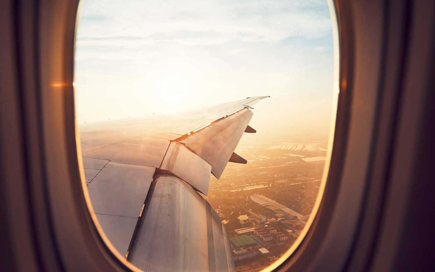 Airplane Window 桌布 壁紙 — 夕陽餘暉灑在飛機機翼上，透過圓形窗戶俯瞰下方廣闊的城市景觀。