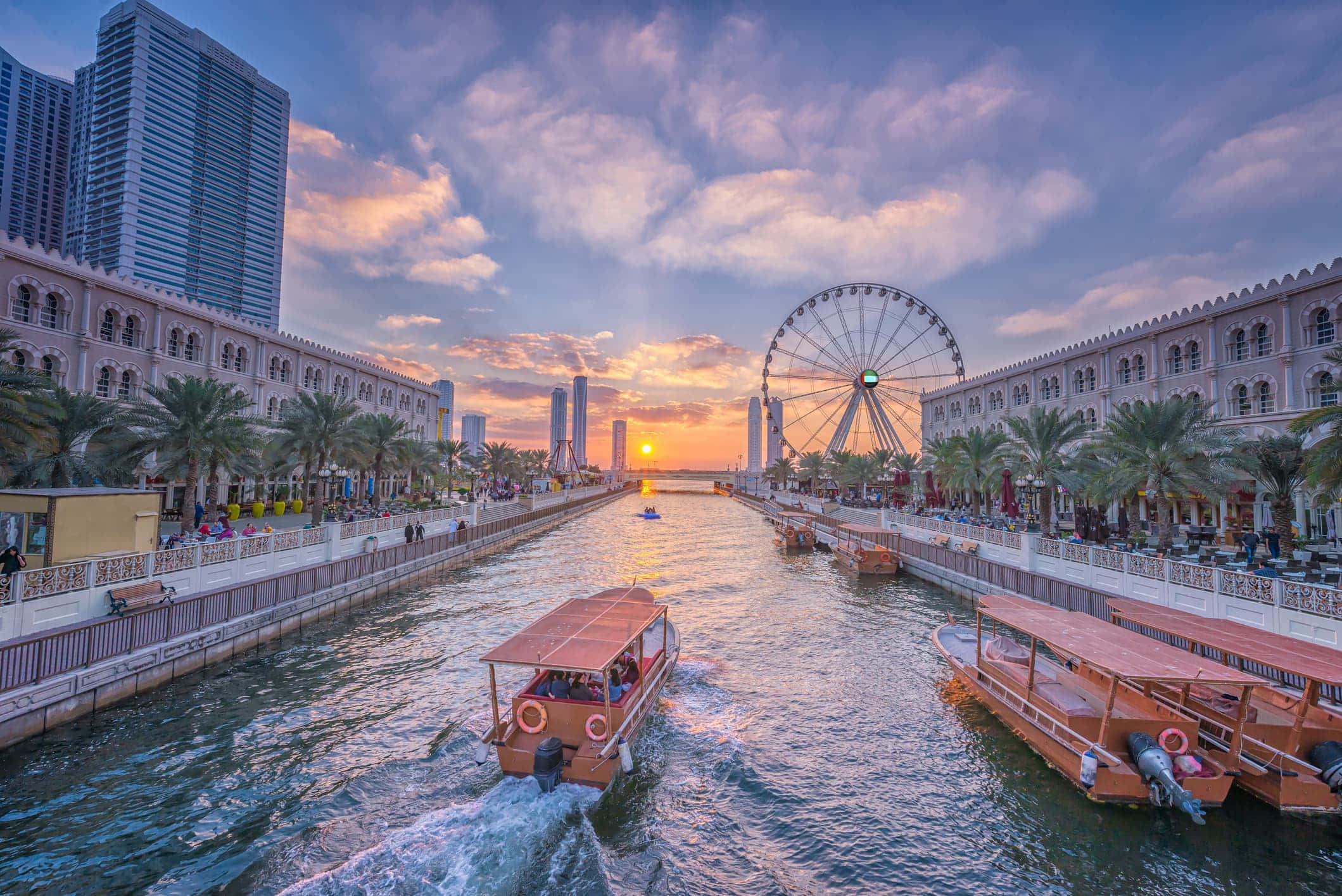 Travel Desktop 桌布 壁紙 — 夕陽餘暉灑落在運河上，有船隻和摩天輪，周圍環繞著棕櫚樹和現代摩天大樓。