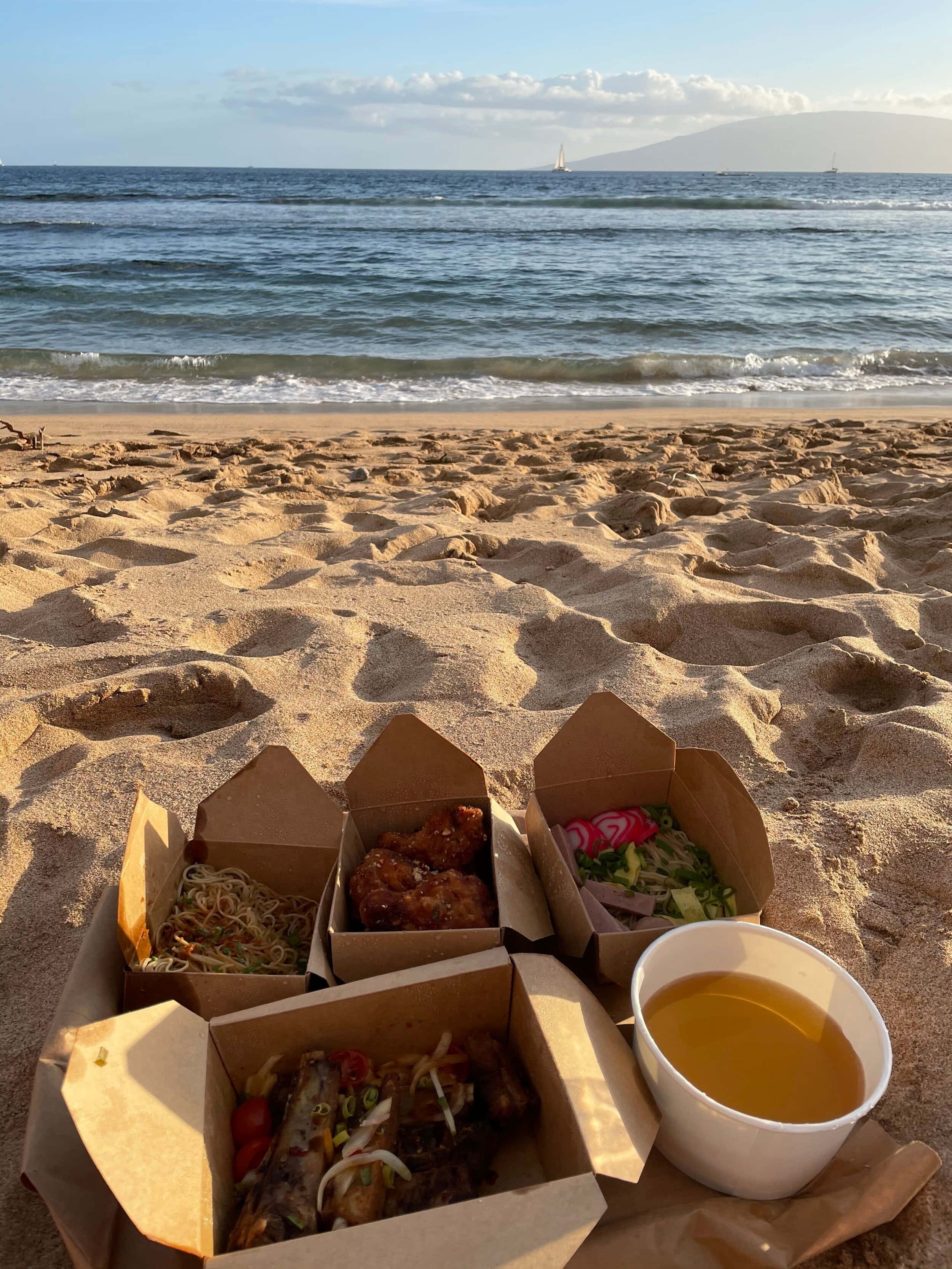 Beach Picnic 桌布 壁紙 — 外帶食物盒和飲料放在沙灘上，背景是海浪和遠處的帆船，天空晴朗。