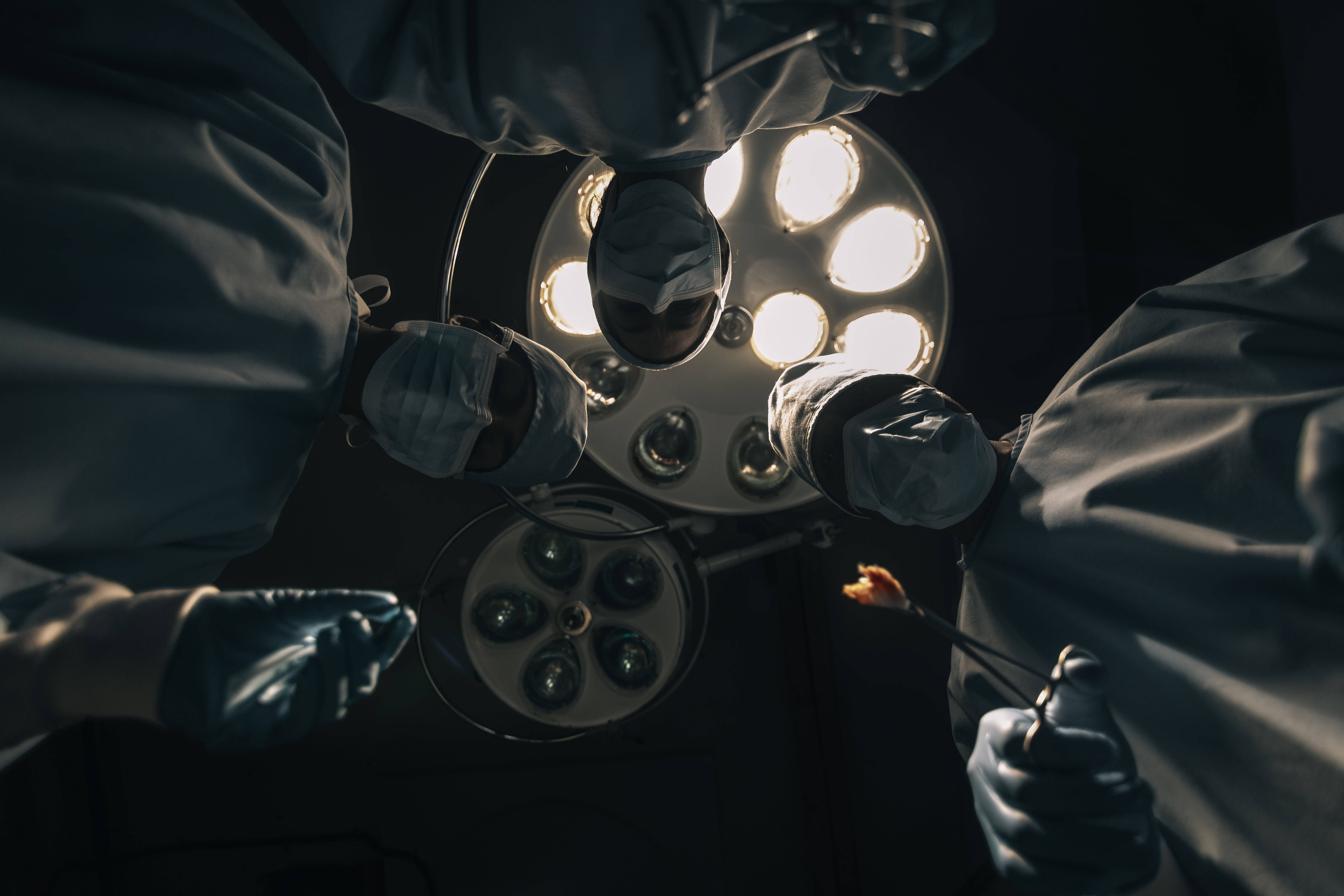 Doctors 桌布 壁紙 — 外科醫生穿著無菌手術衣和口罩，在昏暗的手術室中，於明亮的手術燈下俯身於病人身上。
