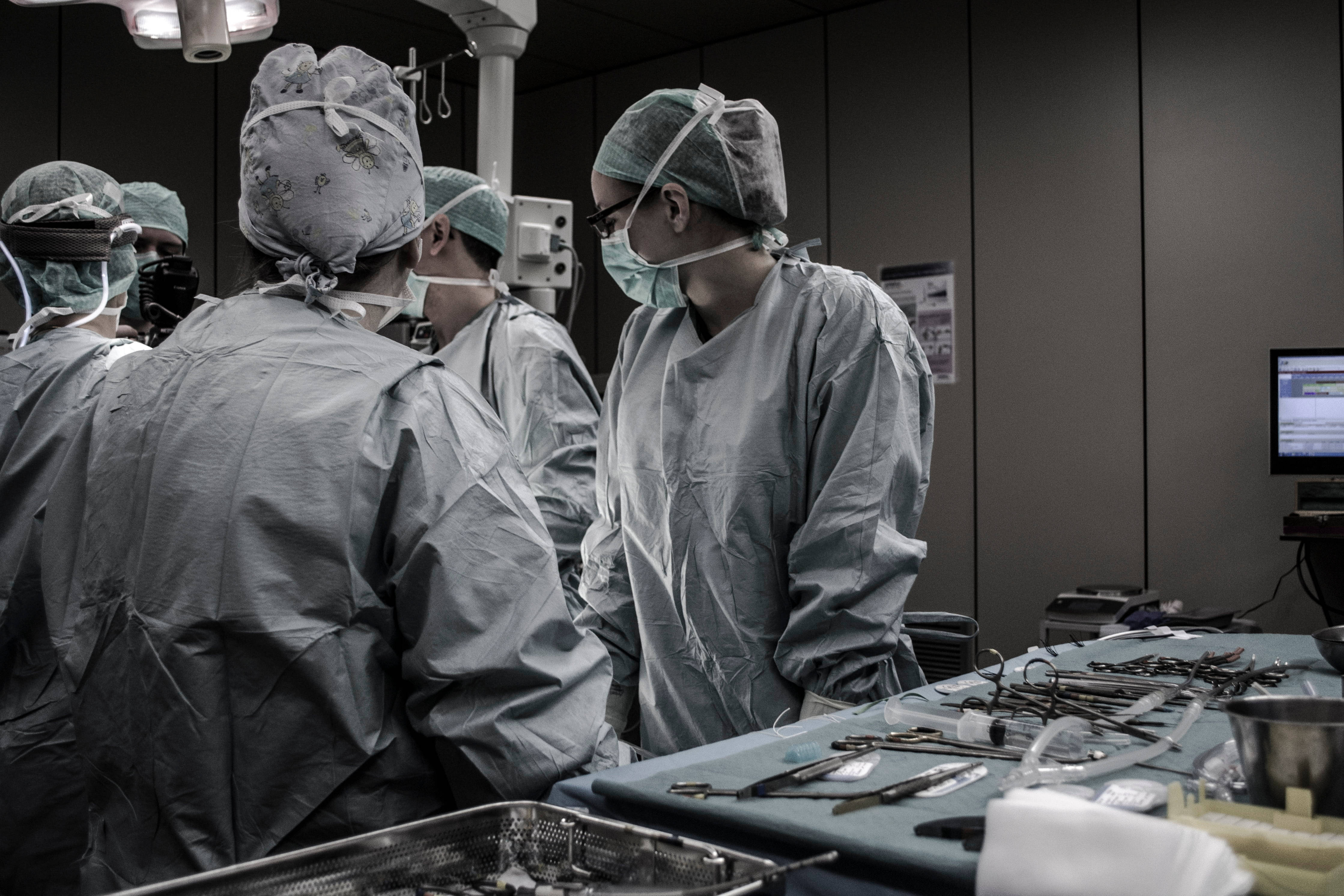 Surgery 桌布 壁紙 — 外科醫生穿著無菌手術袍和口罩，在明亮的頭頂燈光下圍著一張佈滿手術工具的手術檯。