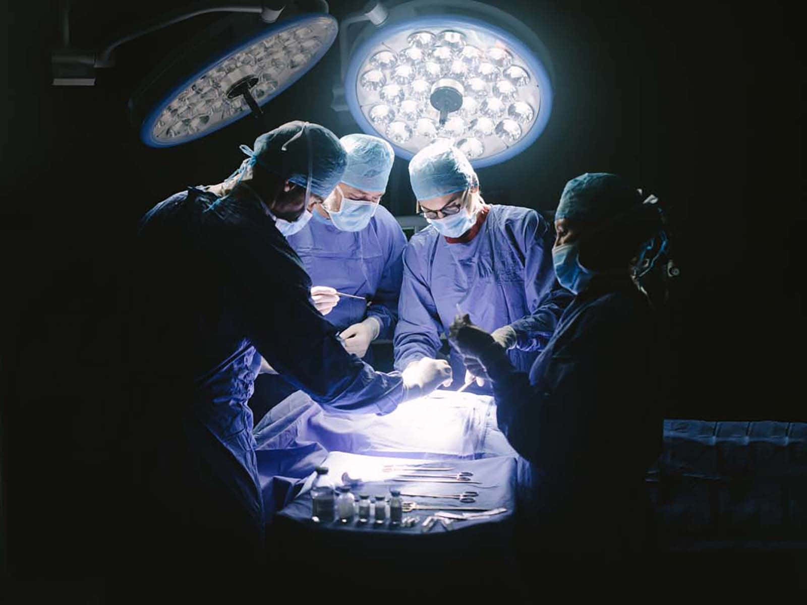 Hospital 桌布 壁紙 — 外科醫生穿著藍色手術服和口罩，在昏暗的手術室中，於明亮的手術燈下專注地進行手術。