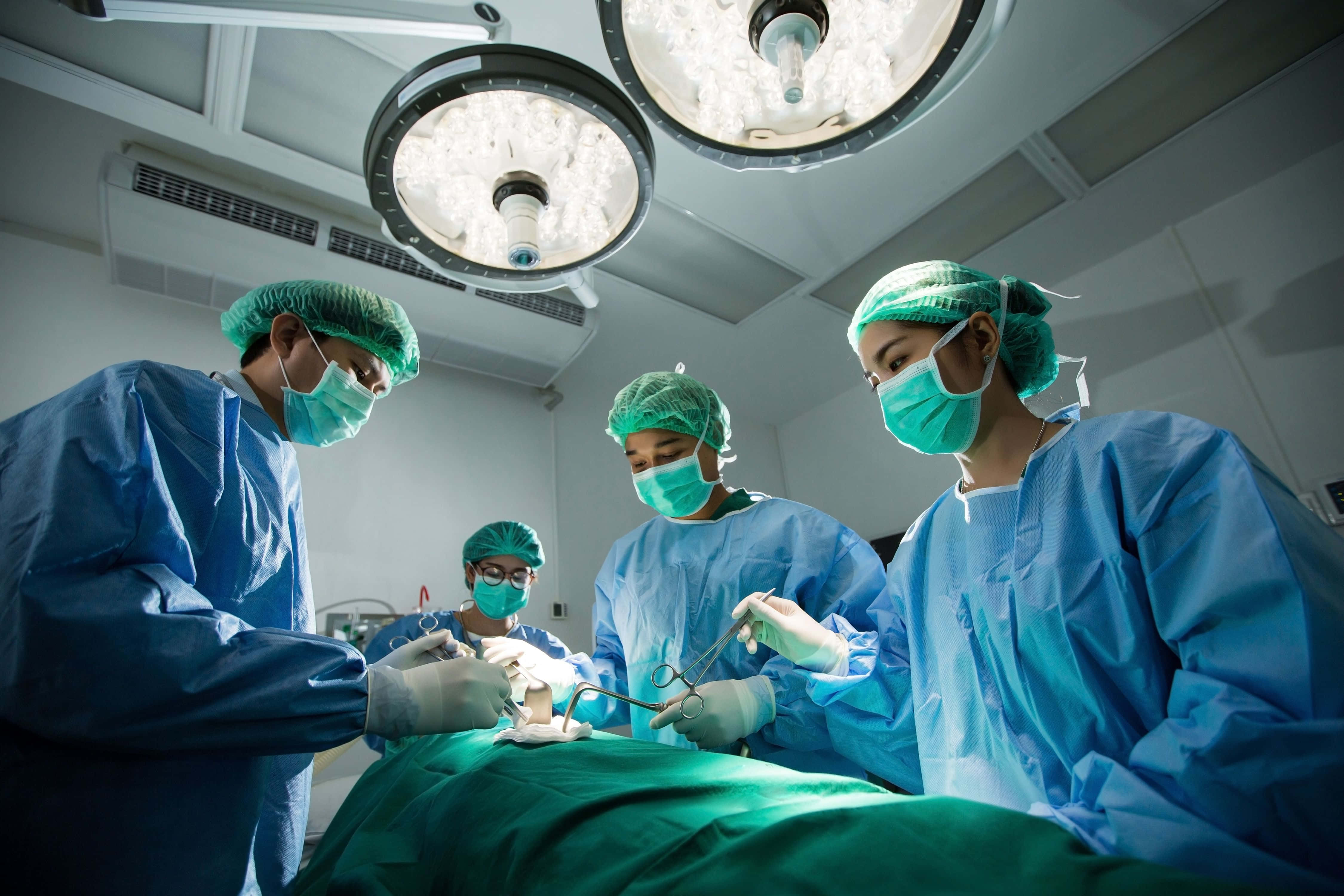 Hospital 桌布 壁紙 — 外科醫生穿著藍色手術服和綠色口罩，在明亮的手術燈下，於無菌手術室中專注地為病人進行手術。