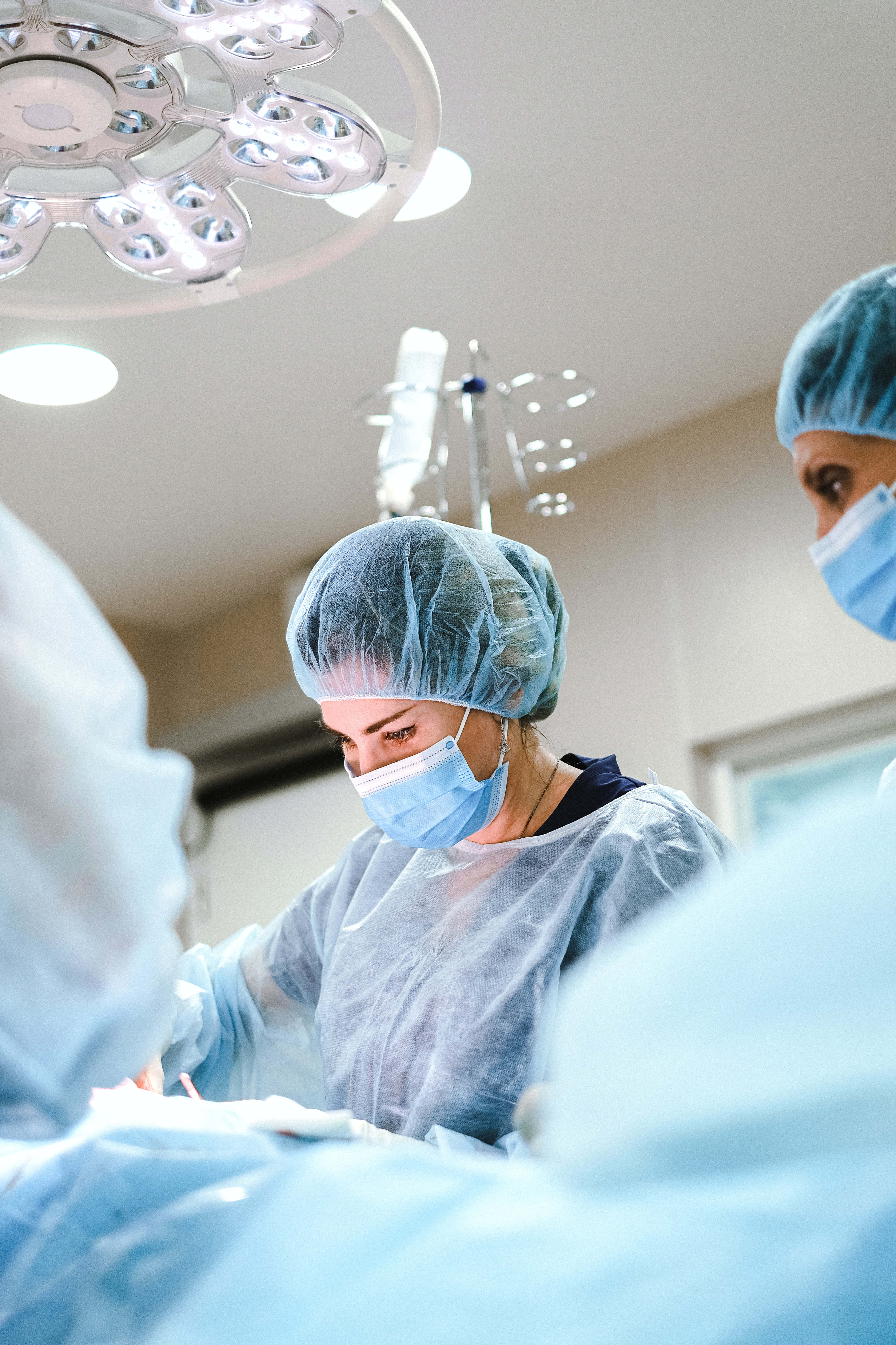 Hospital 桌布 壁紙 — 外科醫生身穿藍色手術衣和口罩，在無菌手術室明亮的無影燈下專注地進行手術。