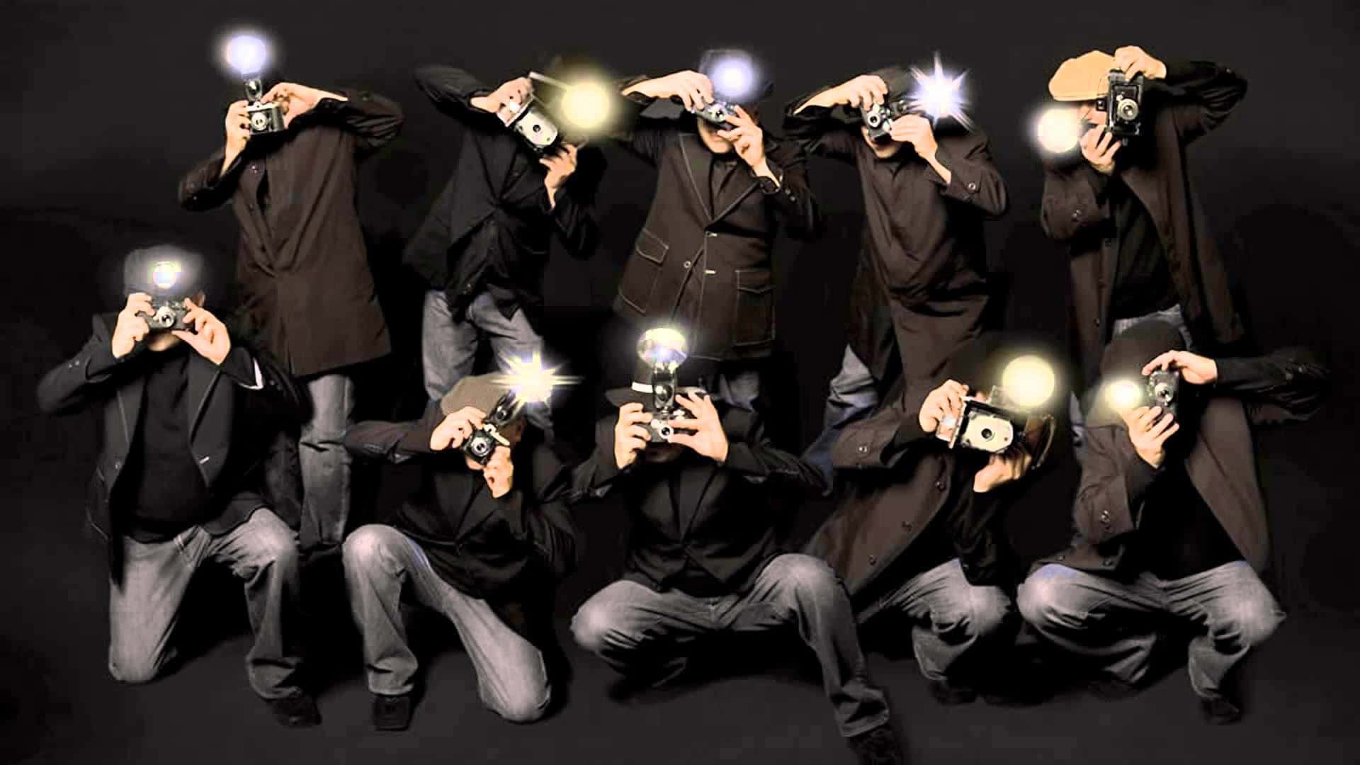 Paparazzi 桌布 壁紙 — 多位身穿深色西裝的攝影師，手持復古相機，在黑色背景下動態擺姿勢，相機發出明亮的閃光。