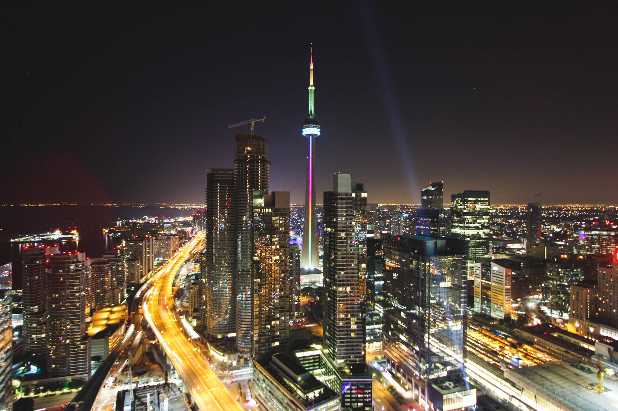 Ontario 桌布 壁紙 — 多倫多令人眼花撩亂的夜景，標誌性的CN塔閃爍著五彩繽紛的燈光，光束穿透黑暗的天空，照耀在由燈火通明的摩天大樓和發光的高速公路組成的網格之上。