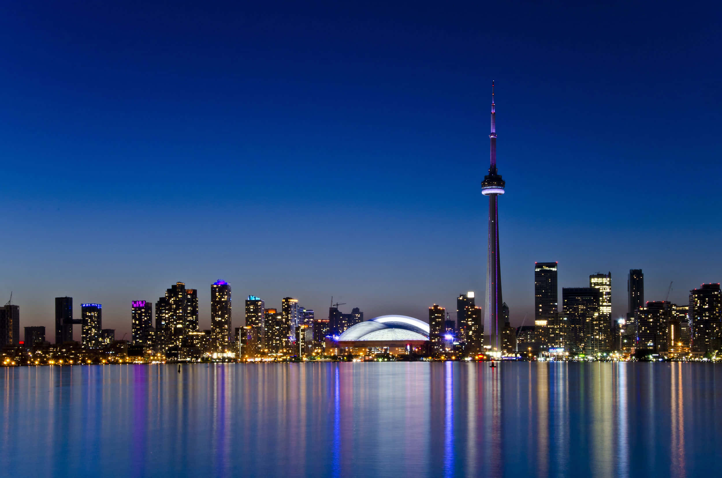 Toronto Skyline 桌布 壁紙 — 多倫多黃昏天際線，CN塔和摩天大樓在深藍色天空下閃耀，倒映在平靜的水面上。
