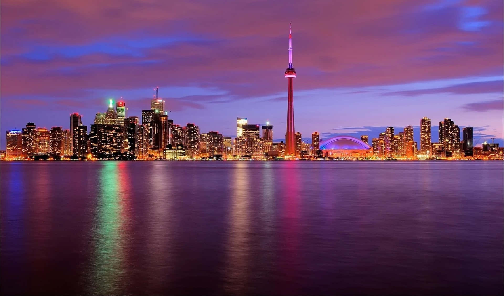 Toronto Skyline 桌布 壁紙 — 多倫多黃昏天際線，CN塔在紫色雲層中閃耀，城市燈光倒映在平靜的水面上。