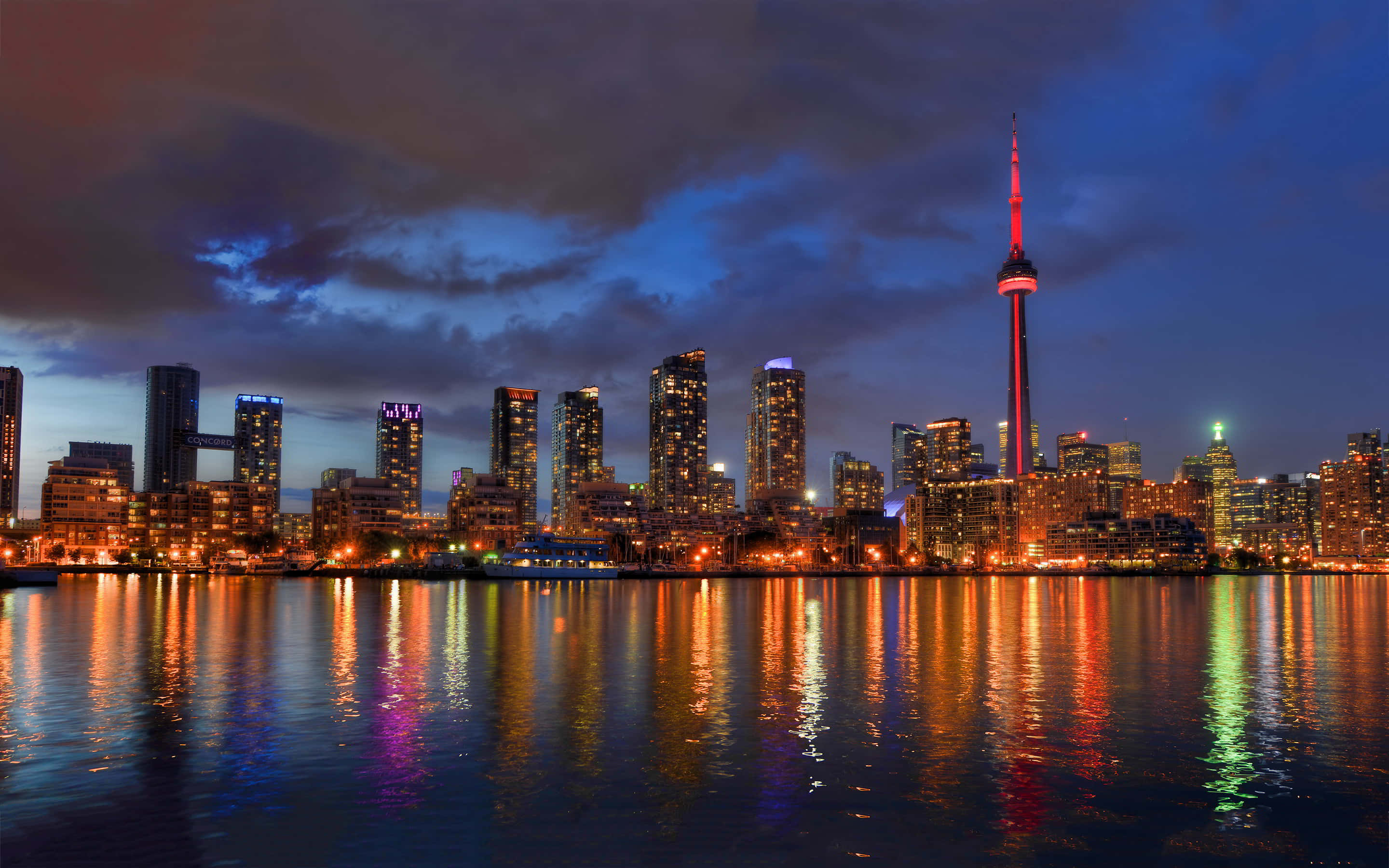 Toronto Skyline 桌布 壁紙 — 多倫多黃昏天際線，CN塔發出紅光，城市燈光在平靜的水面上倒映，天空有戲劇性的雲層。