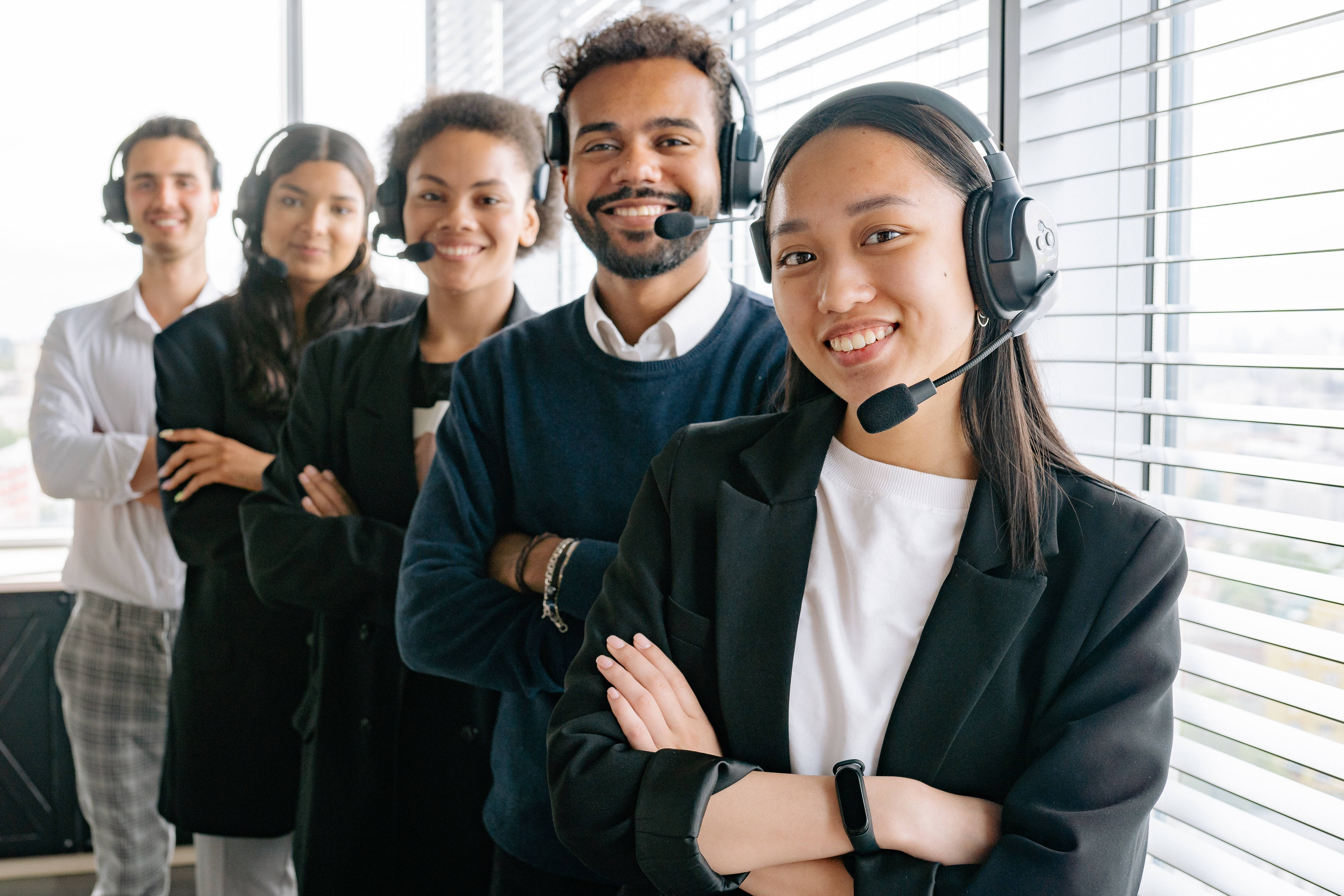 Working 桌布 壁紙 — 多元化的微笑客戶服務團隊戴著耳機，自信地站在帶有百葉窗的明亮現代辦公室中。