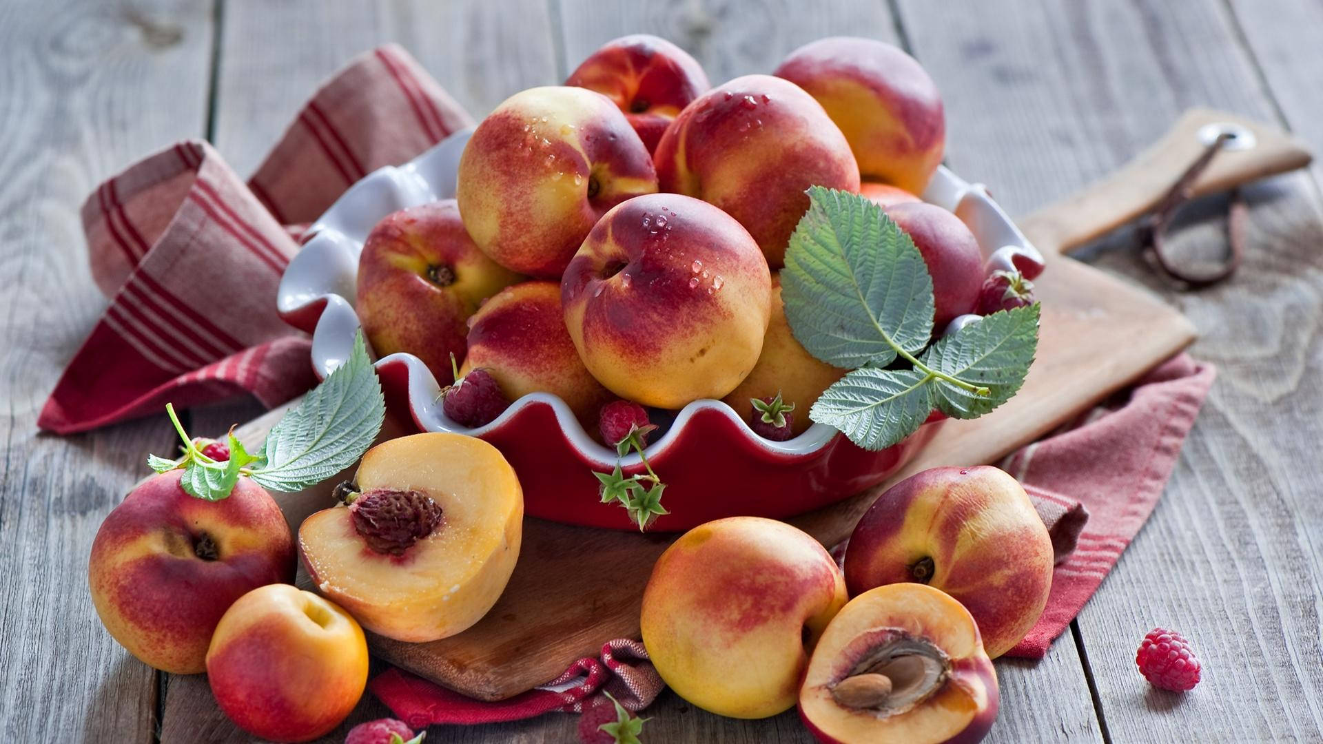 Fruits 桌布 壁紙 — 多汁成熟的桃子和覆盆子從一個紅色陶瓷碗中溢出，碗放在鋪有條紋布的鄉村風格木桌上。