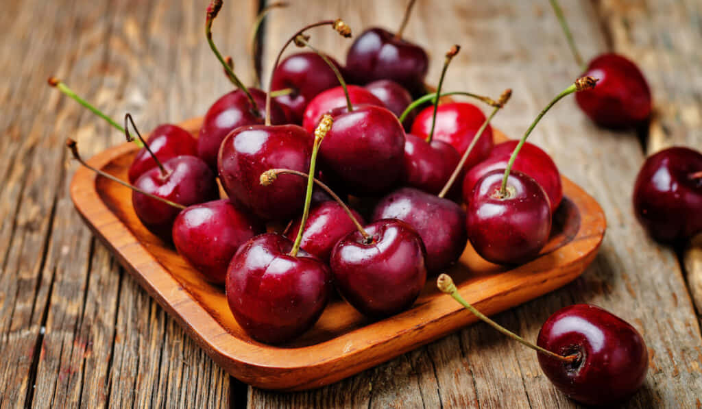Red Fruit 桌布 壁紙 — 多汁成熟的櫻桃連莖在質樸木盤上閃閃發光，置於經風化的木桌上。