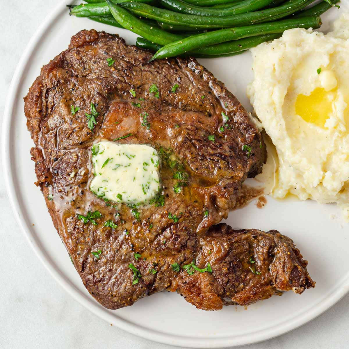 Ribeye Steak 桌布 壁紙 — 多汁烤牛排配上香草奶油，旁邊有馬鈴薯泥和青豆，放在白色盤子上。