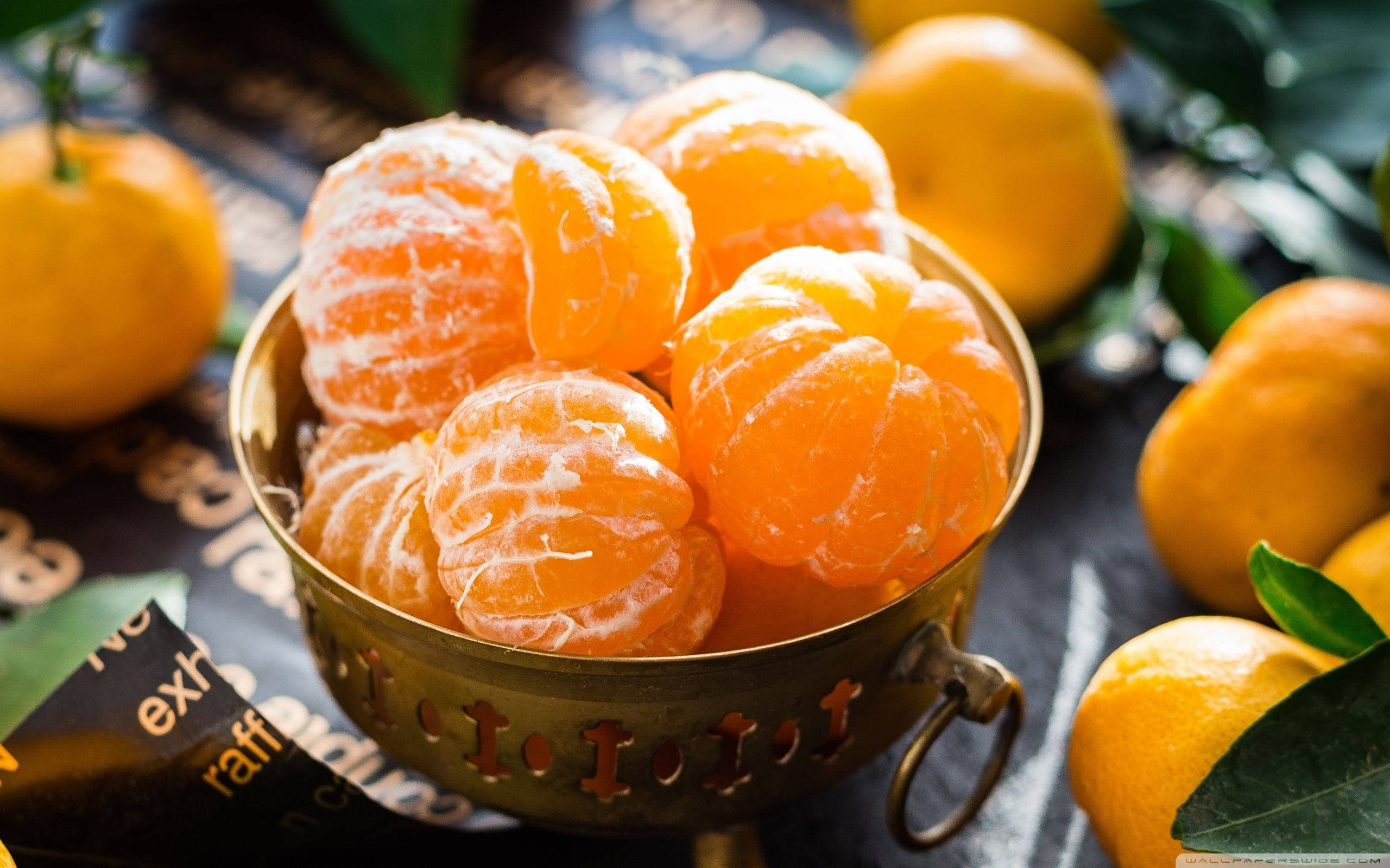 Fruits 桌布 壁紙 — 多汁的剝皮柑橘在復古黃銅碗中閃閃發光，周圍環繞著完整的水果和綠葉，置於深色圖案表面上。