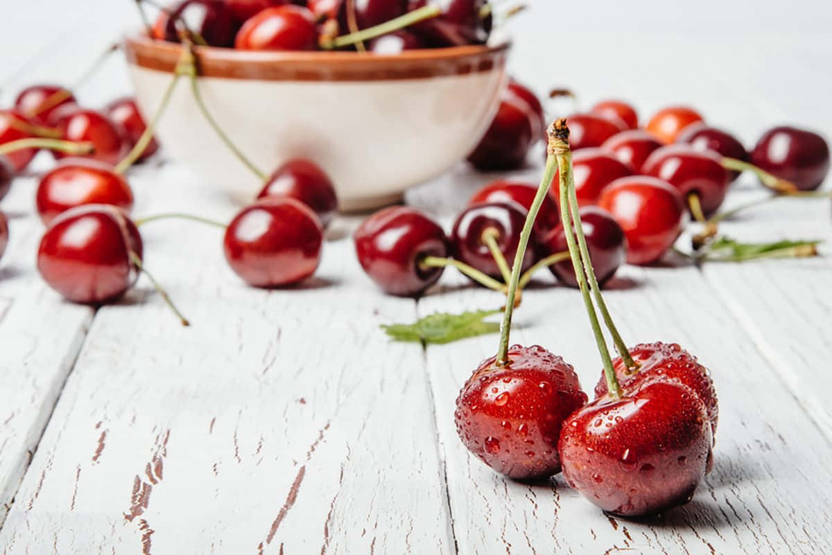 Red Cherries 桌布 壁紙 — 多汁的紅櫻桃在白色木質表面上閃爍著露珠，後面有一個柔焦的陶瓷碗。