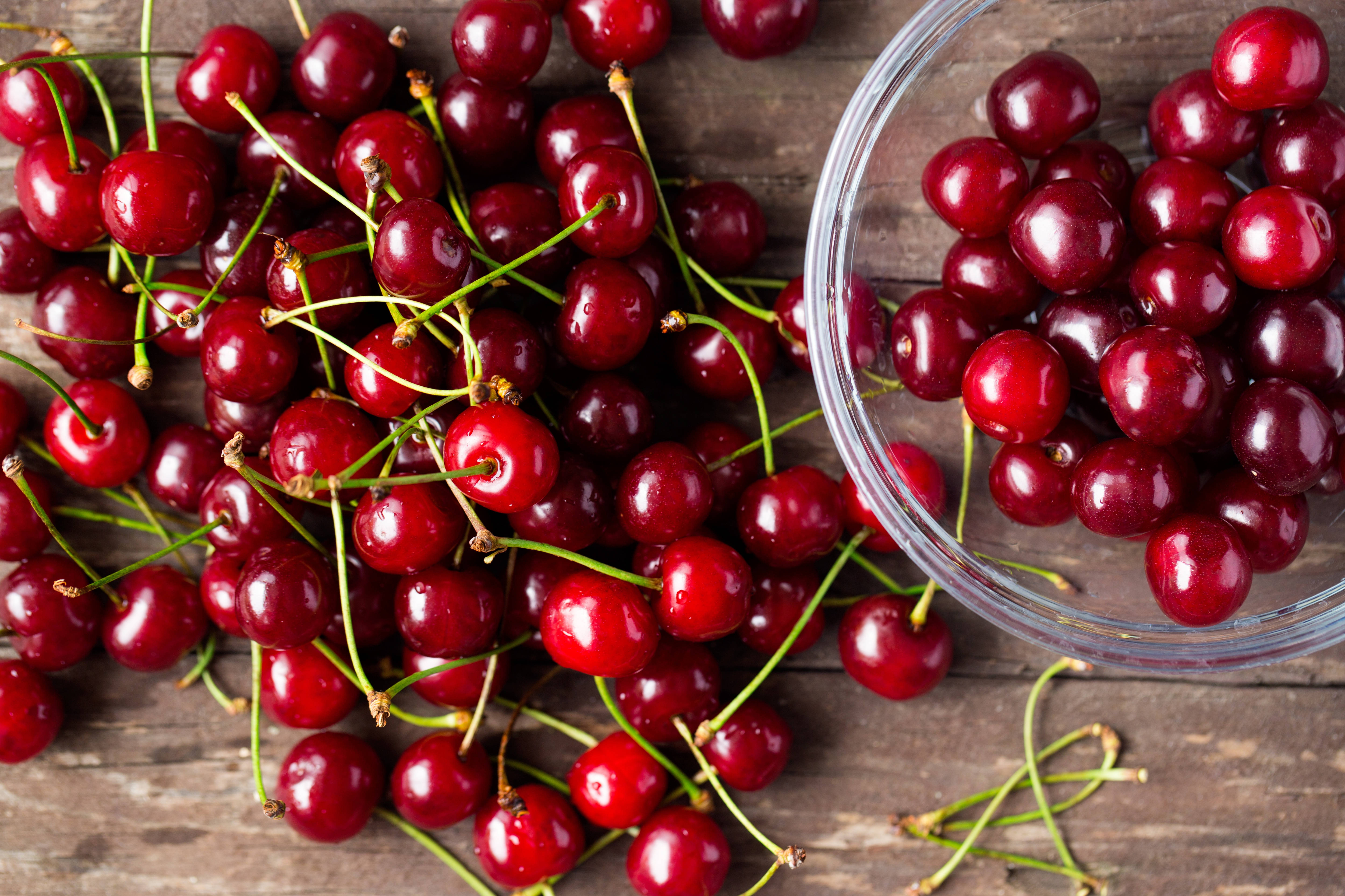 Cherry 桌布 壁紙 — 多汁的紅櫻桃帶著綠色莖從玻璃碗中溢出，落在質樸的木質表面上。