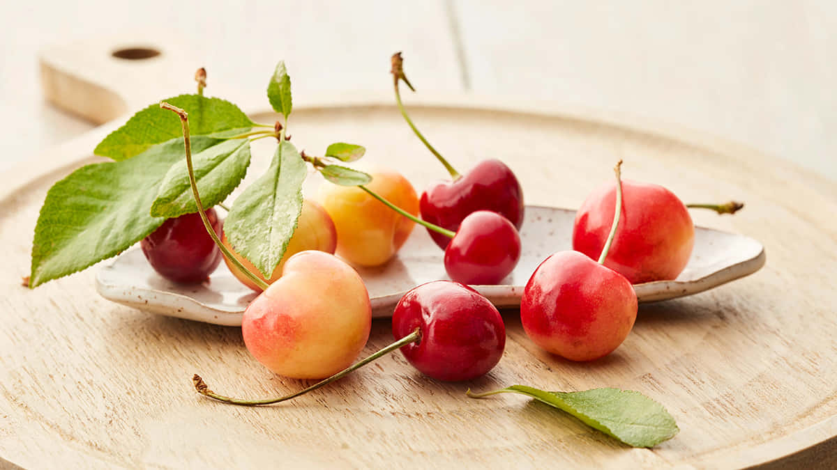 Red Cherries 桌布 壁紙 — 多汁的紅色和黃色櫻桃，配上綠葉，放在淺色木質表面上的陶瓷盤上。