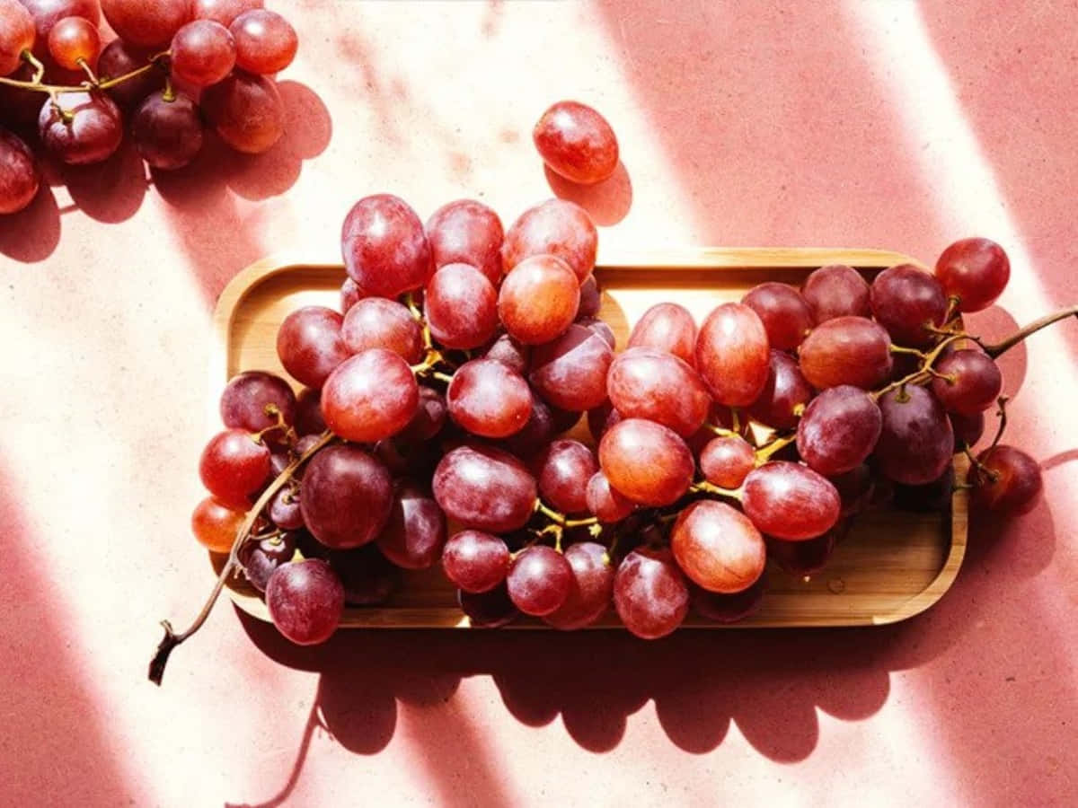 Grape 桌布 壁紙 — 多汁的紅葡萄在溫暖的陽光下閃閃發光，放在木托盤上，背景是柔軟的粉紅色紋理。