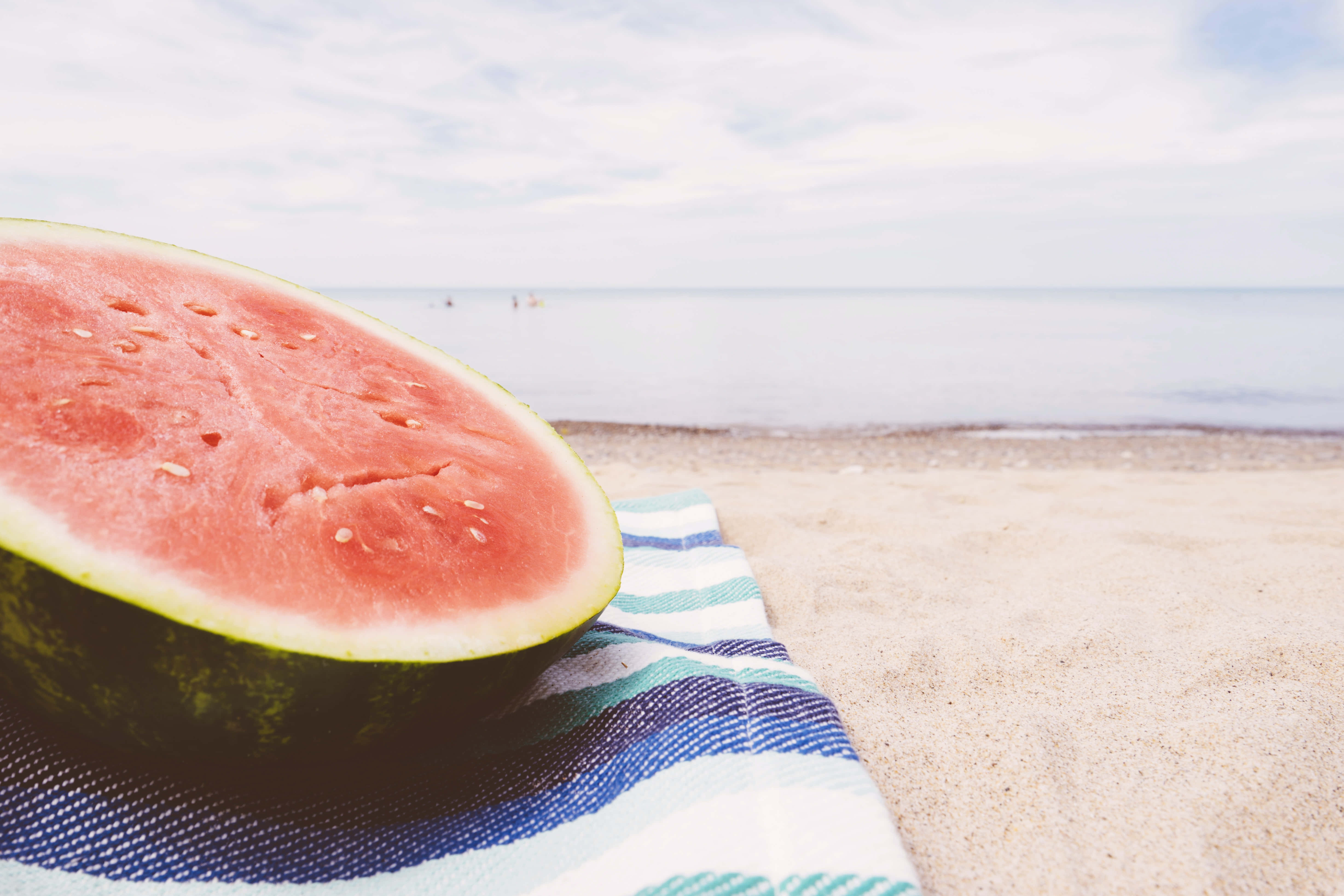 Summer Fruits 桌布 壁紙 — 多汁的西瓜半個放在沙灘上的條紋毛巾上，背景是平靜的海洋和多雲的天空，柔和焦點。