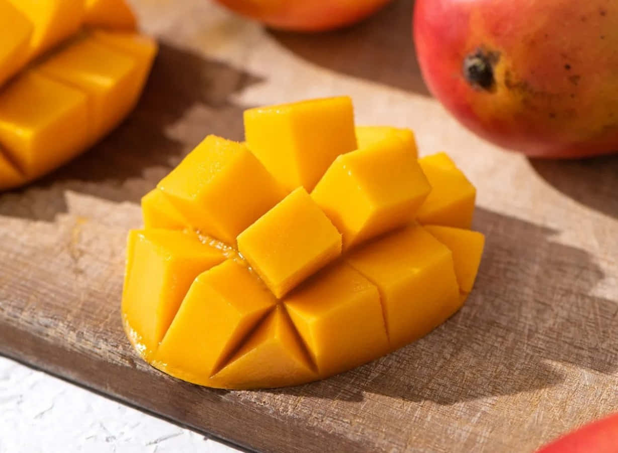 Mango 桌布 壁紙 — 多汁的金色芒果丁在質樸的木砧板上排列成網格狀，背景有軟焦點的完整芒果。