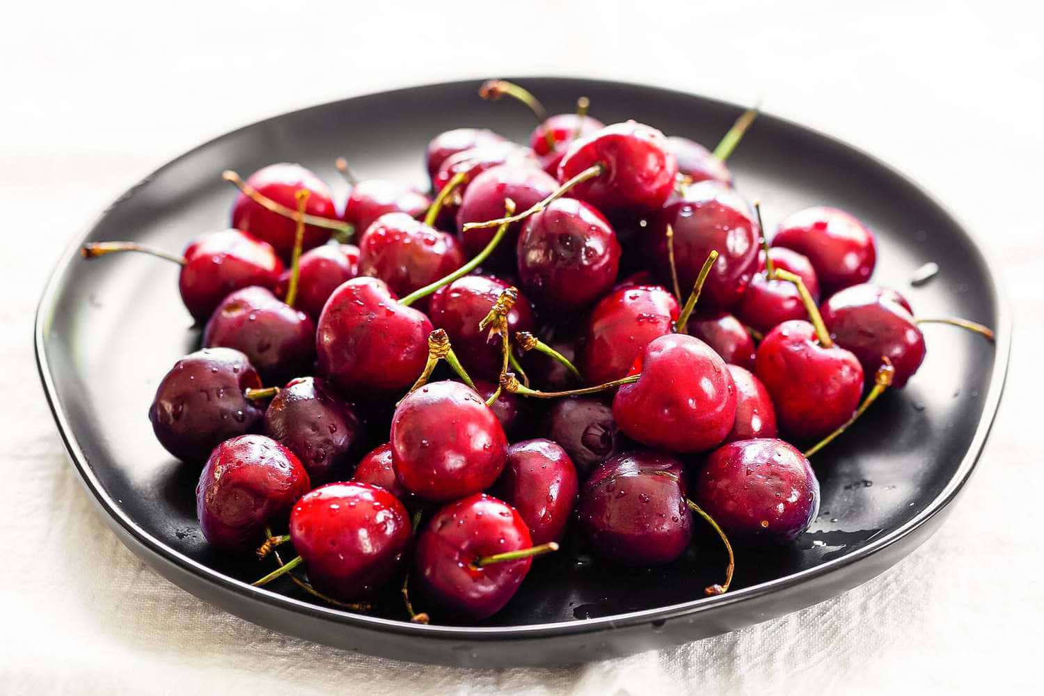 Red Cherries 桌布 壁紙 — 多汁飽滿的櫻桃閃爍著水滴，高高堆疊在霧面黑色盤子上，背景是柔軟的白色紋理。