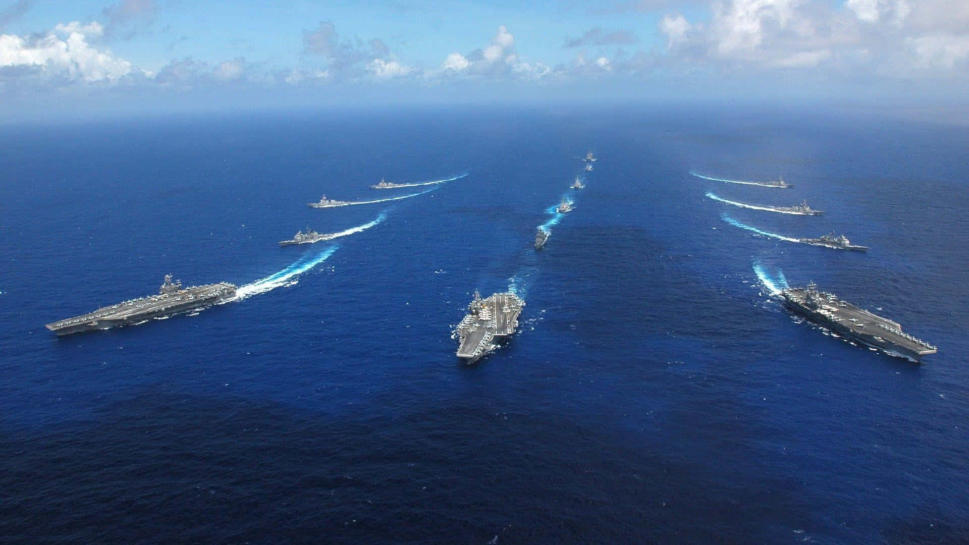 Navy 桌布 壁紙 — 多艘航空母艦和軍艦在部分多雲的天空下，於深藍色海洋上編隊巡航的鳥瞰圖。