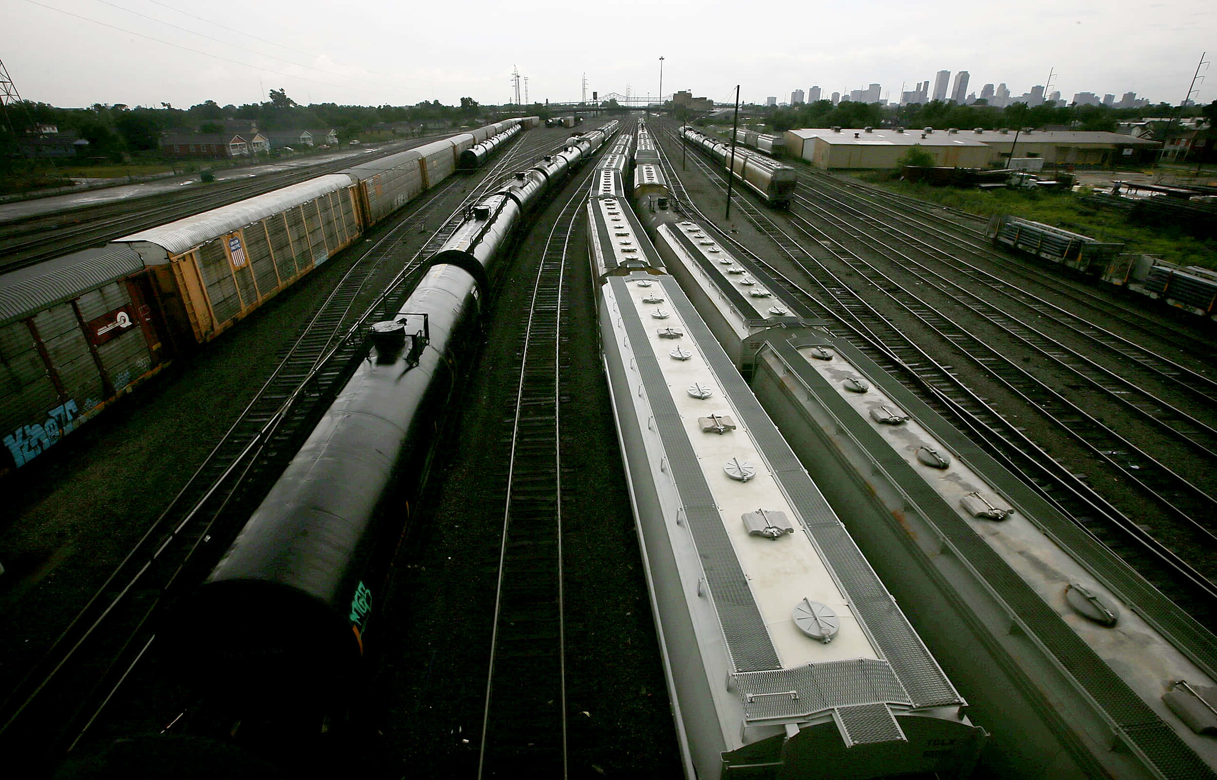 Train Tracks 桌布 壁紙 — 多輛貨運列車在工業鐵路調車場的平行軌道上，遠方城市天際線在陰天下的空拍視圖。