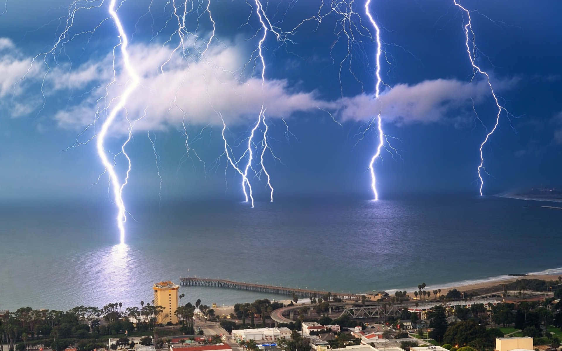 Thunderstorm 桌布 壁紙 — 多道明亮的閃電擊中沿海城鎮上方的海洋，天空是暴風雨的藍色。