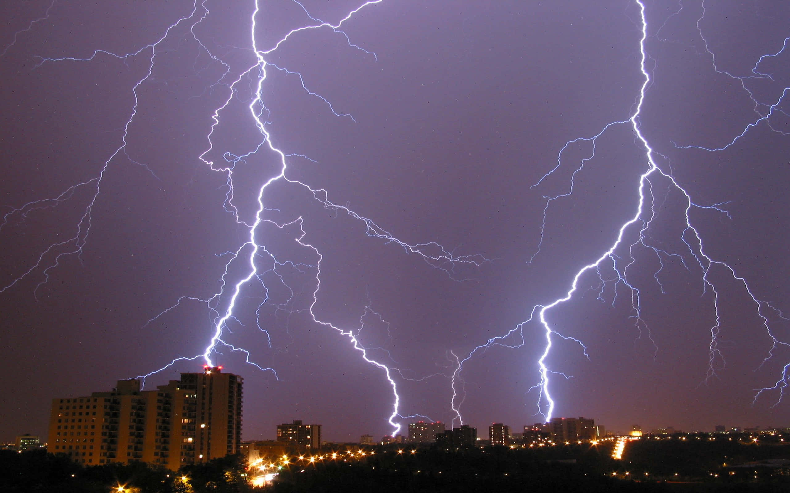 閃電 桌布 壁紙 — 多道鋸齒狀閃電照亮了暴風雨夜晚的天空，下方是燈火通明的城市景觀。