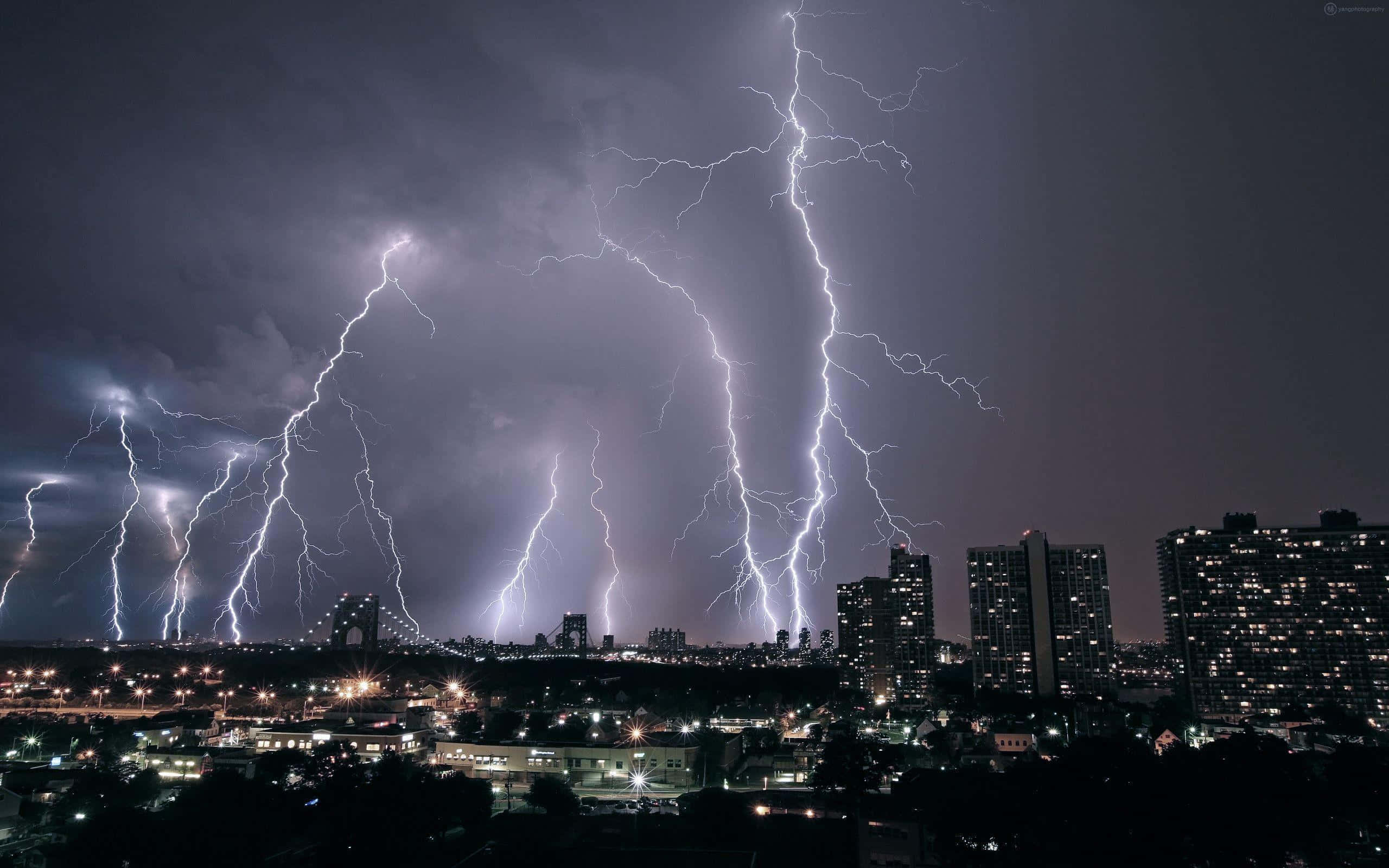 閃電 桌布 壁紙 — 多道鋸齒狀閃電照亮了暴風雨的夜空，下方是城市景觀，有亮燈的建築物和遠處的橋樑。