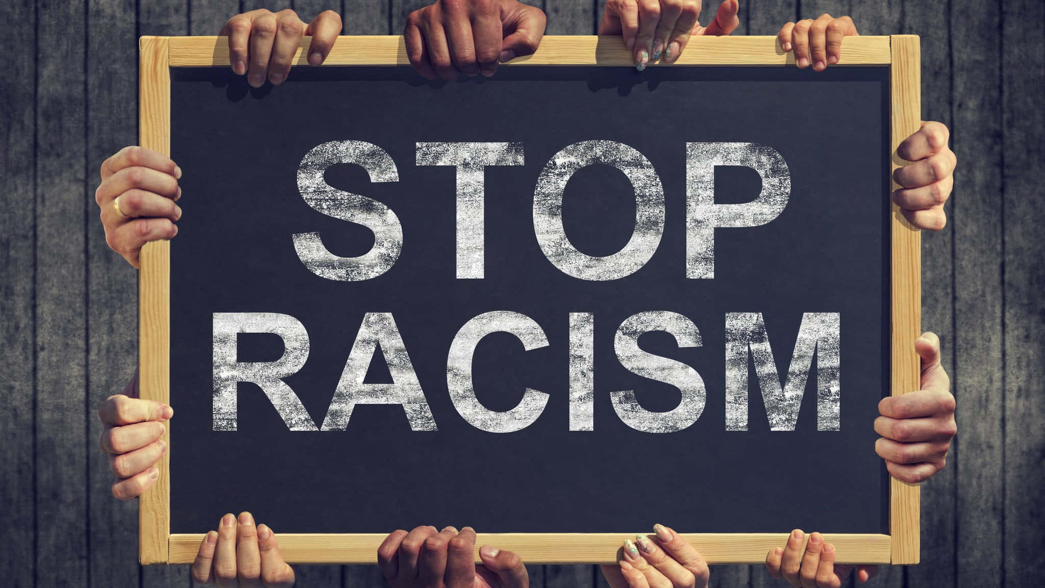 stop 桌布 壁紙 — 多隻手舉起一個木框黑板，上面以粗體顯示「STOP RACISM」的訊息，背景是風化的木牆。