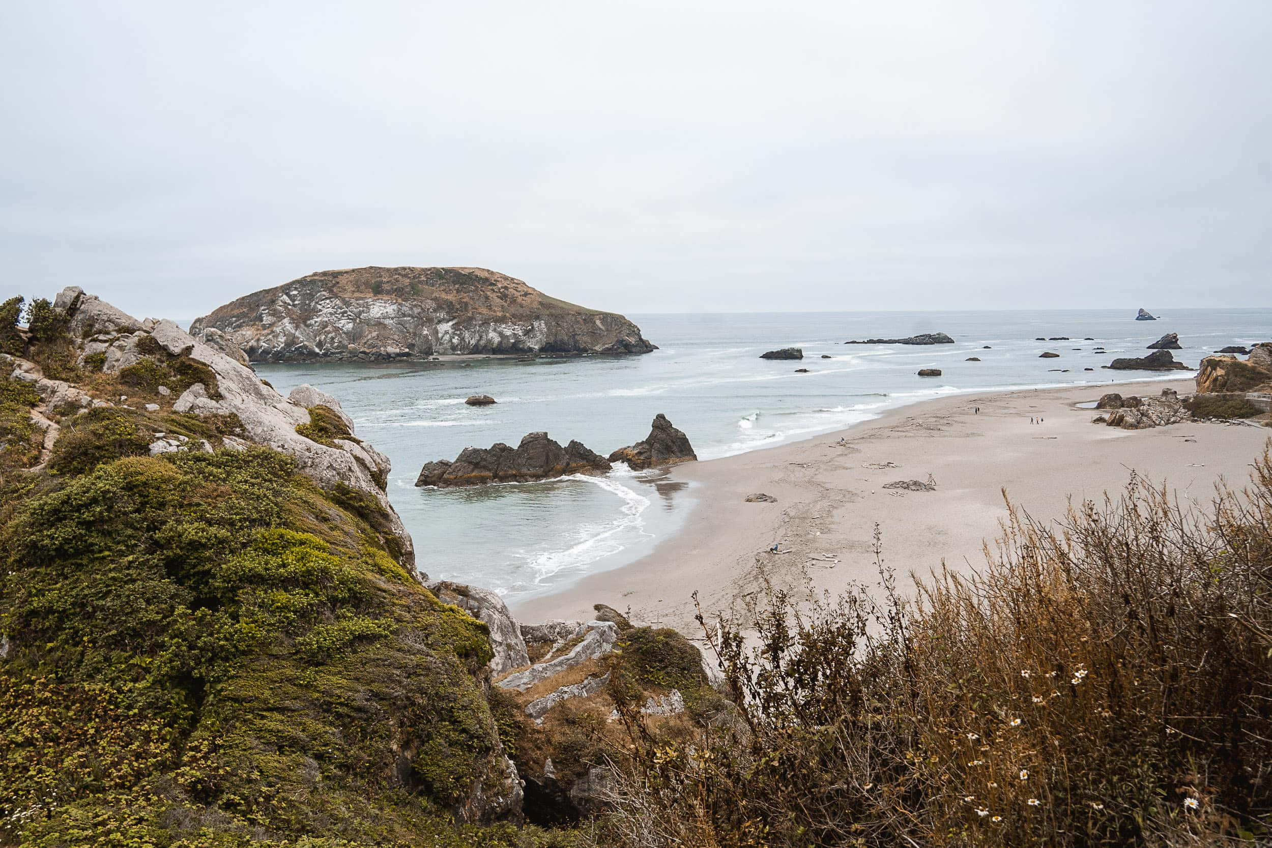 Oregon Coast 桌布 壁紙 — 多雲天空下，一個霧氣瀰漫的海岸場景，沙灘繞著岩石露頭和遠處島嶼彎曲。