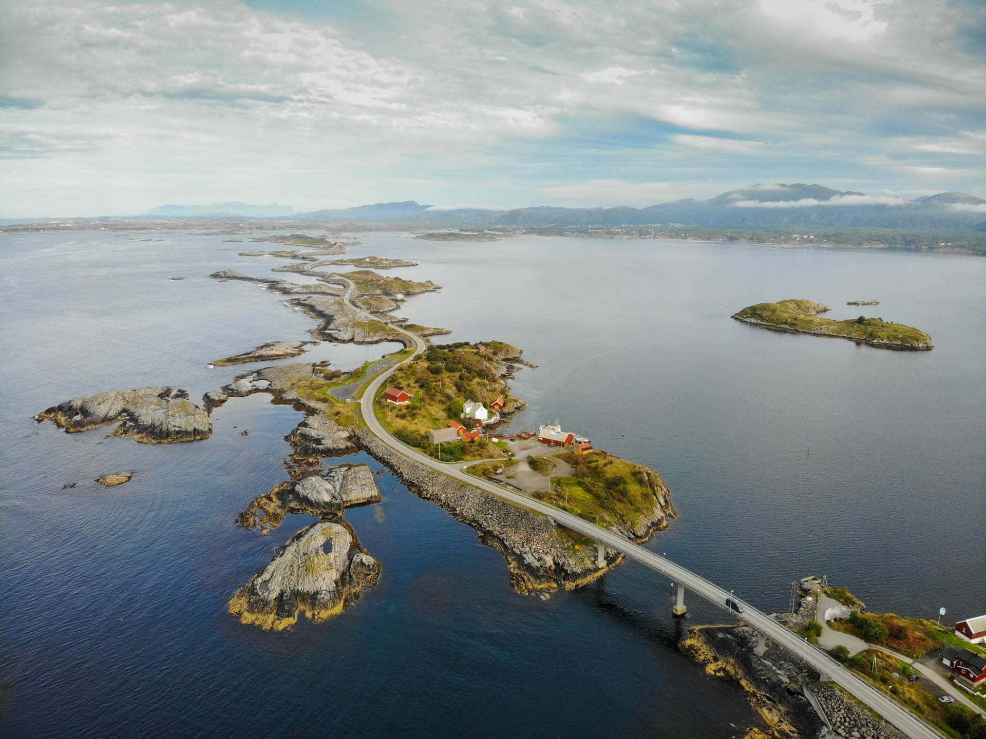 Road 桌布 壁紙 — 多雲天空下，一條蜿蜒的海岸道路連接岩石島嶼上紅頂房屋的鳥瞰圖。