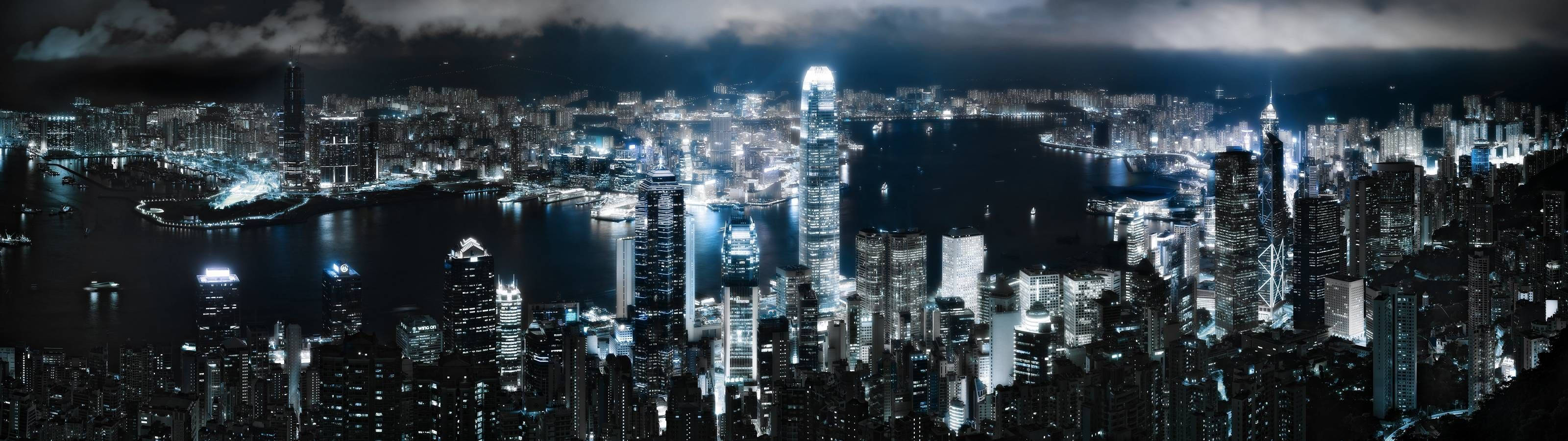 Dual 桌布 壁紙 — 多雲天空下，密集燈光的城市景觀全景夜景，高聳的摩天大樓倒映在黑暗的水道上。