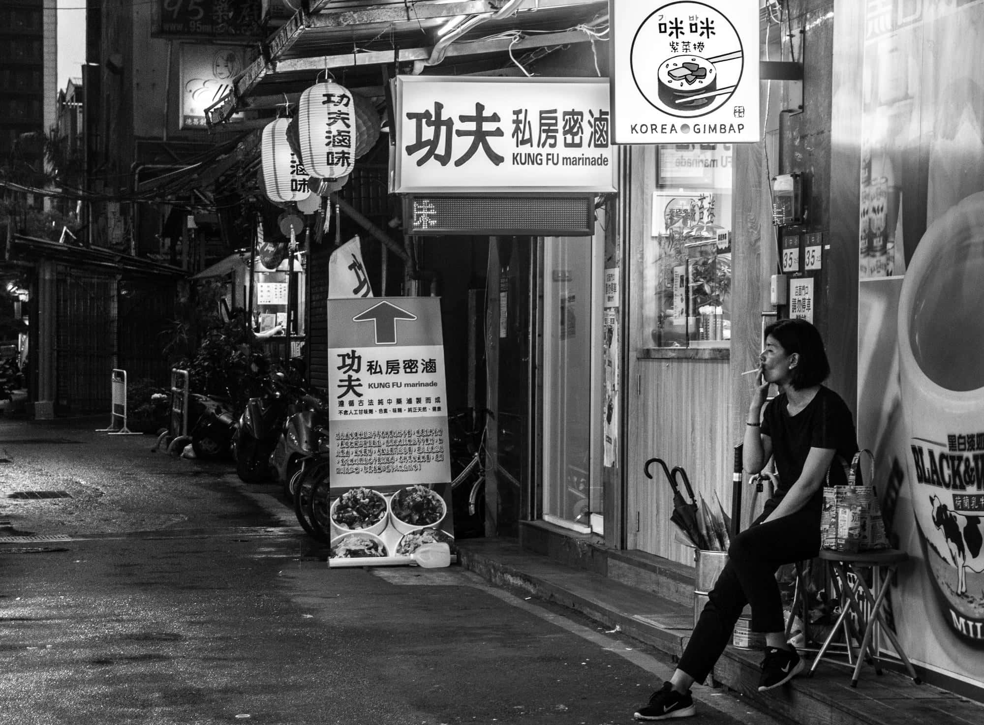 日本黑白 桌布 壁紙 — 夜晚，一位女士獨自坐在狹窄小巷中韓國餐廳外的凳子上，周圍掛著招牌和停放的機車。