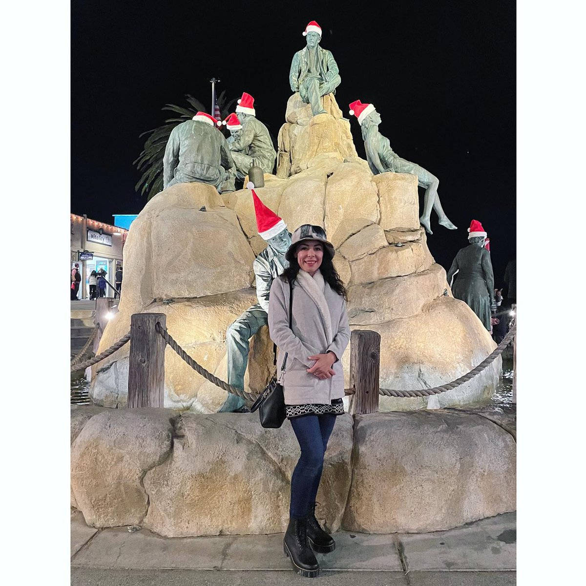 Woman 桌布 壁紙 — 夜晚，一位戴帽子的微笑女士站在一組戴著紅色聖誕帽的節日雕像前。