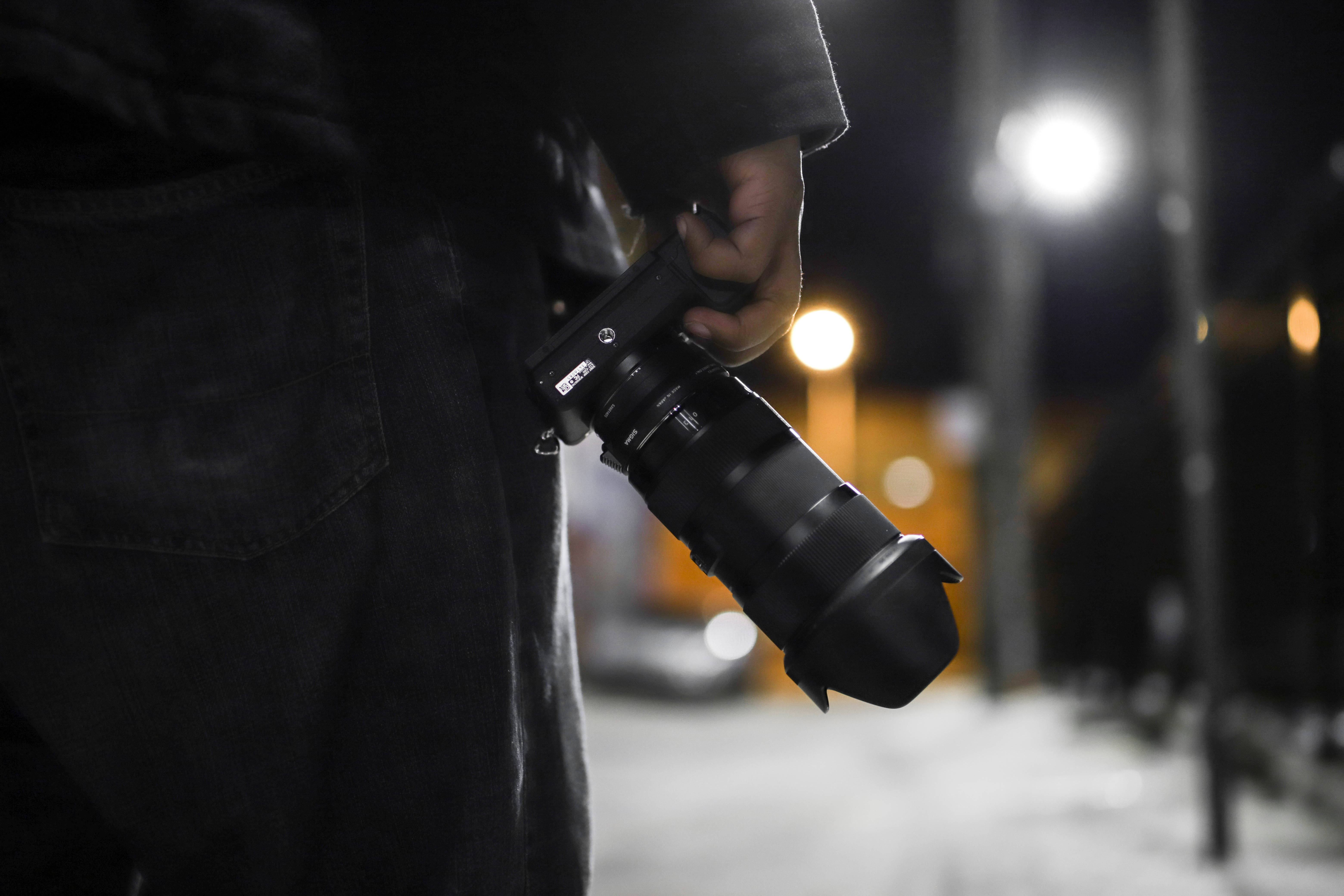 Sony 桌布 壁紙 — 夜晚，一個人拿著一個大相機鏡頭，背景中模糊的路燈柔和地發光。
