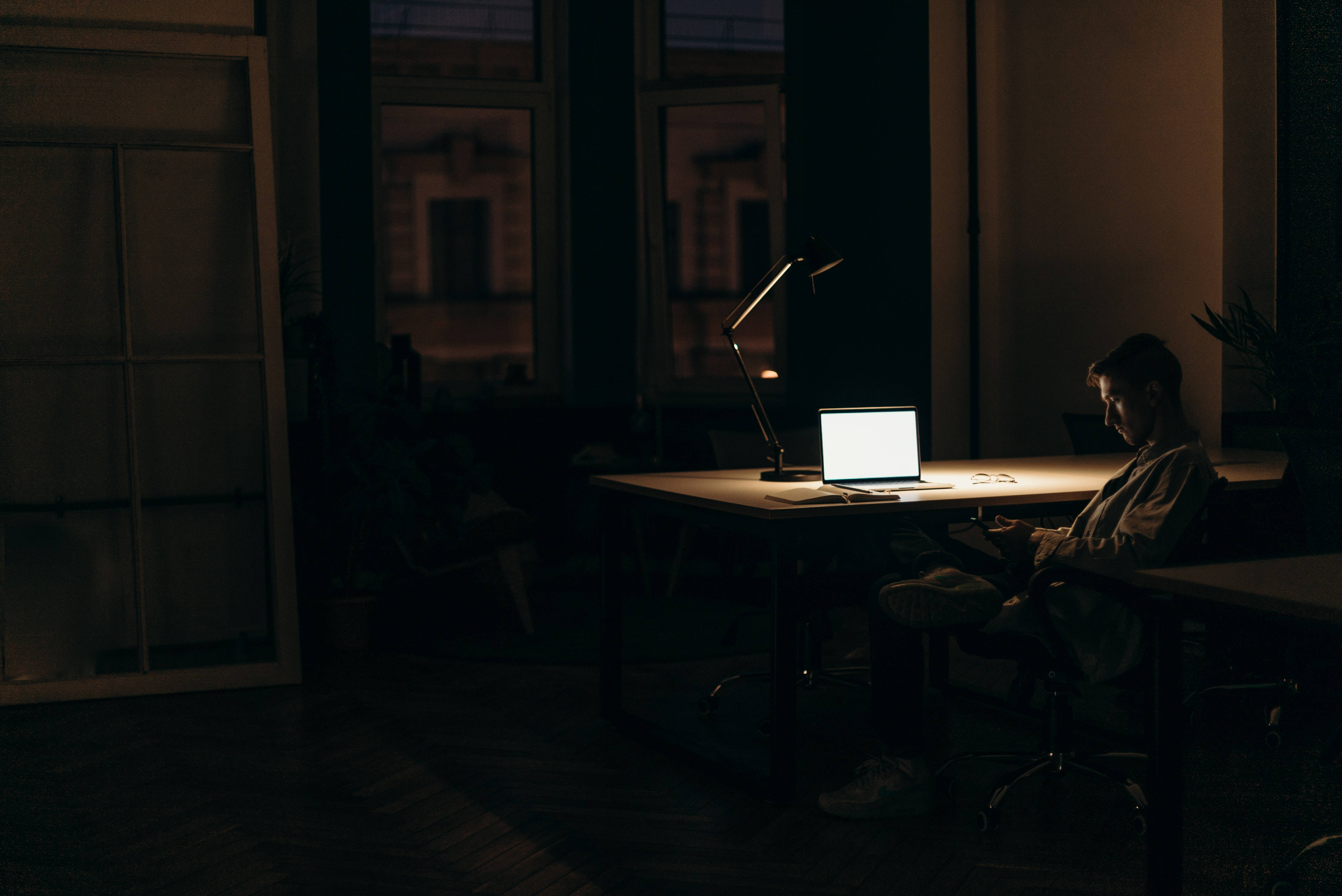 Work 桌布 壁紙 — 夜晚，一個人獨自坐在昏暗的房間裡，被筆記型電腦和檯燈照亮的書桌前。