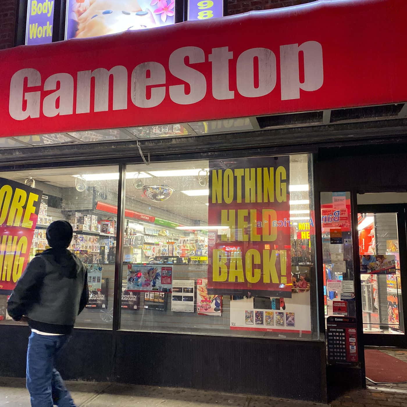 GameStop 桌布 壁紙 — 夜晚，一個人走過一間燈火通明的GameStop商店，窗戶上有醒目的招牌和促銷海報。