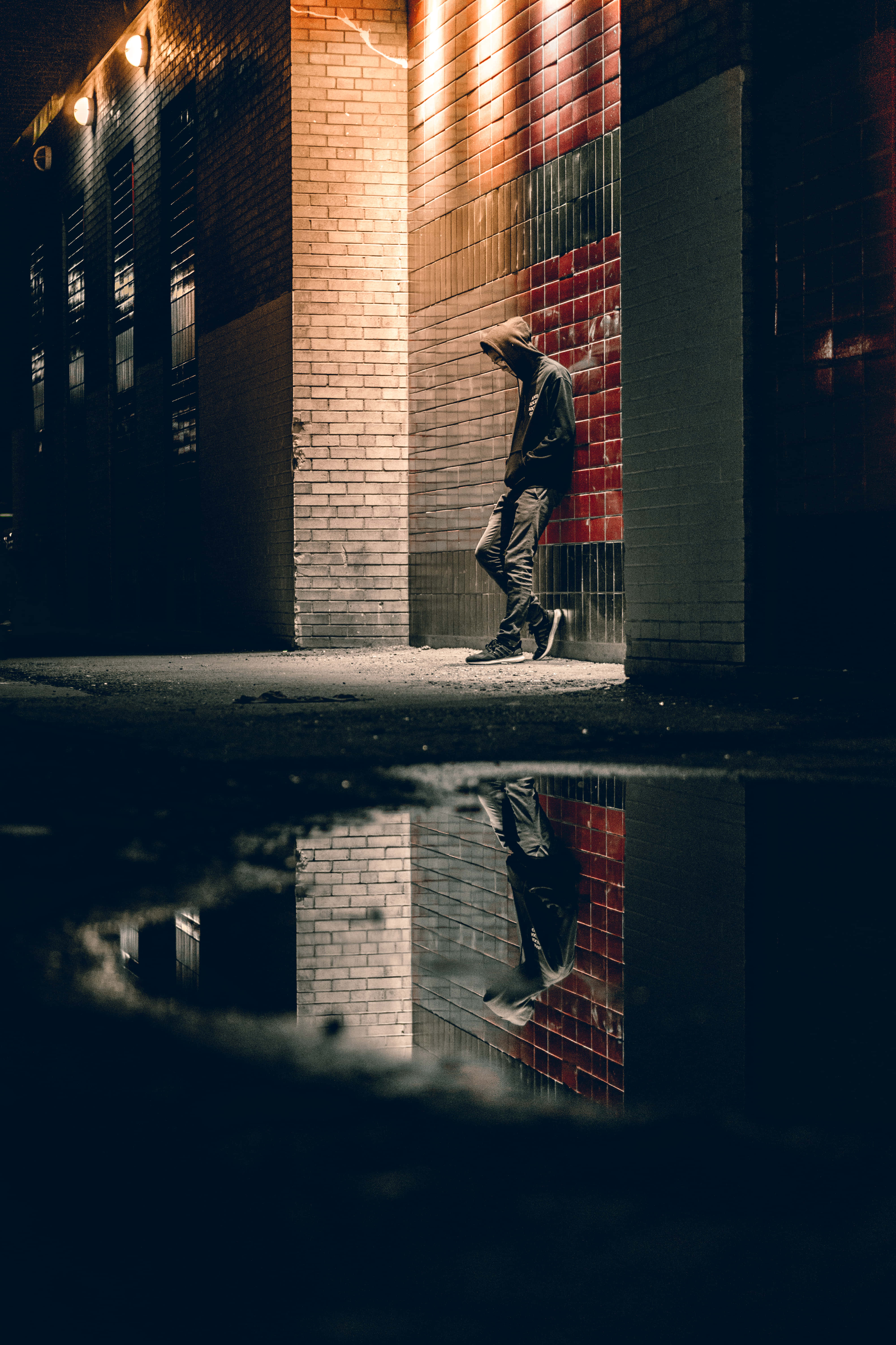 Sad Face 桌布 壁紙 — 夜晚，一個孤獨的戴帽人影靠在磚牆上，他們的倒影映照在下面的雨水坑中。