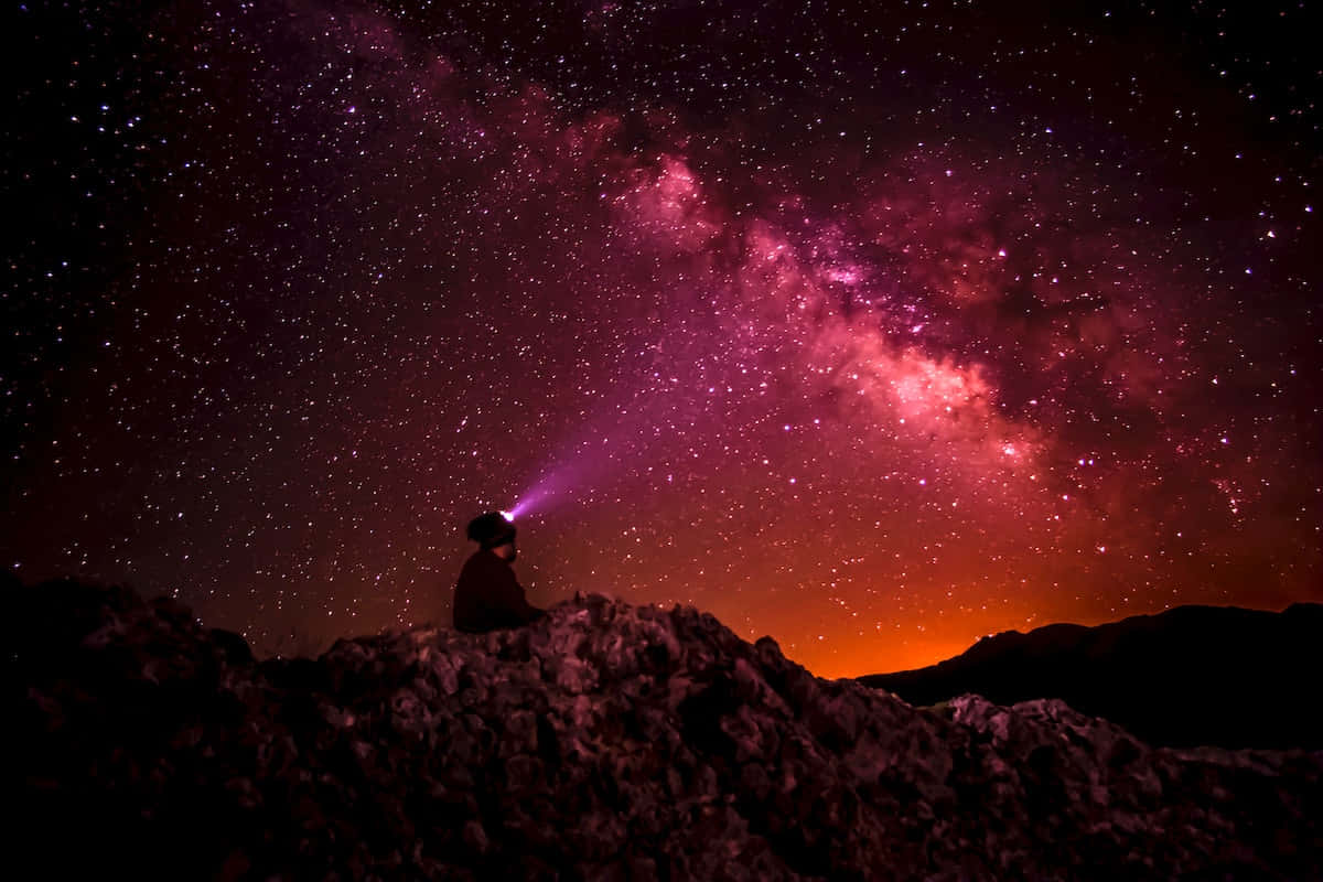 Beautiful Galaxy 桌布 壁紙 — 夜晚，一個孤獨的身影坐在岩石山脊上，用頭燈照向充滿活力的粉紅色和橙色銀河系。