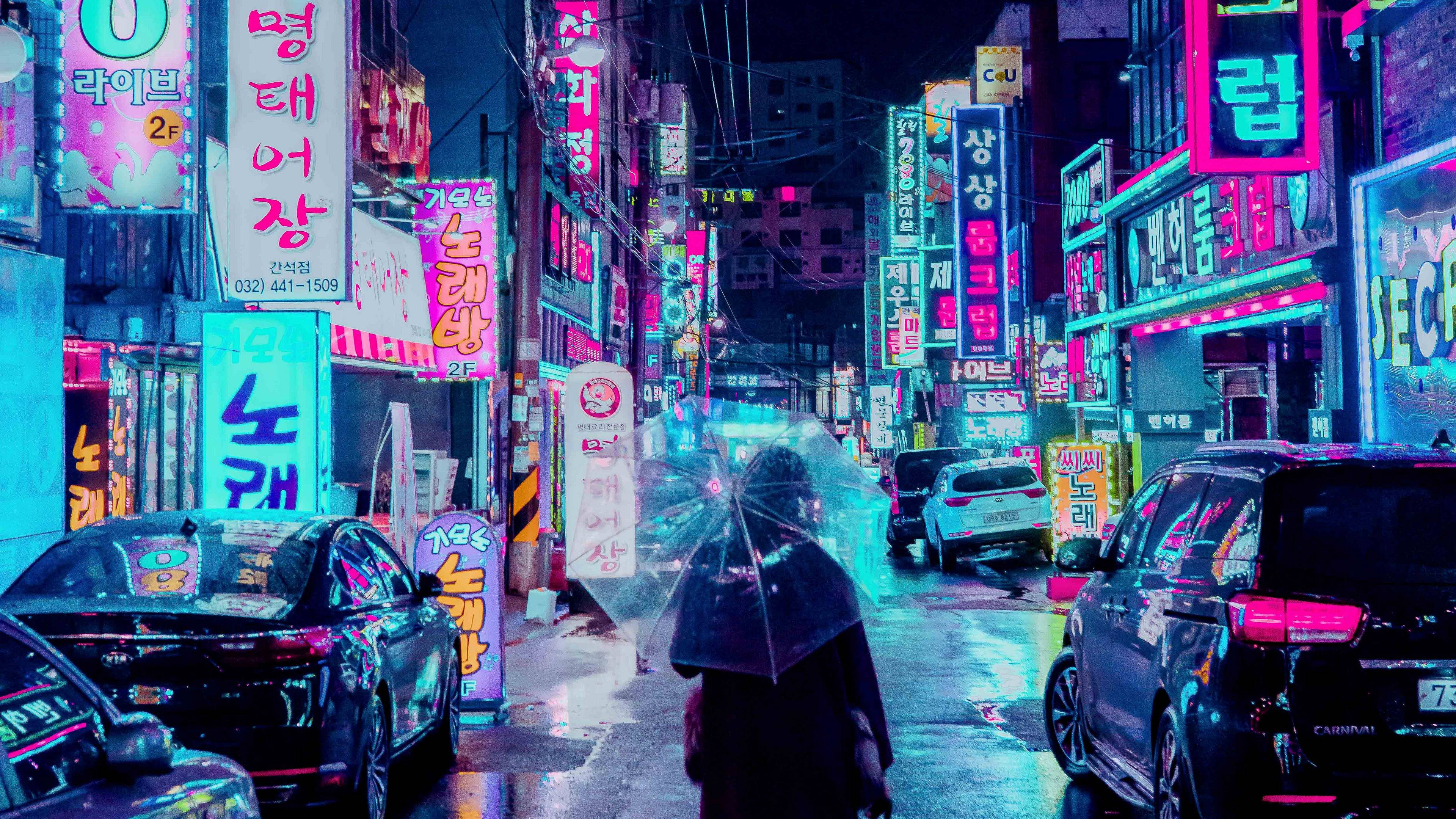 桌布 壁紙 — 夜晚，一個孤獨的身影撐著傘走過一條充滿霓虹燈的韓國小巷，雨水在潮濕的路面上反射出鮮豔的粉紅色、藍色和綠色招牌。