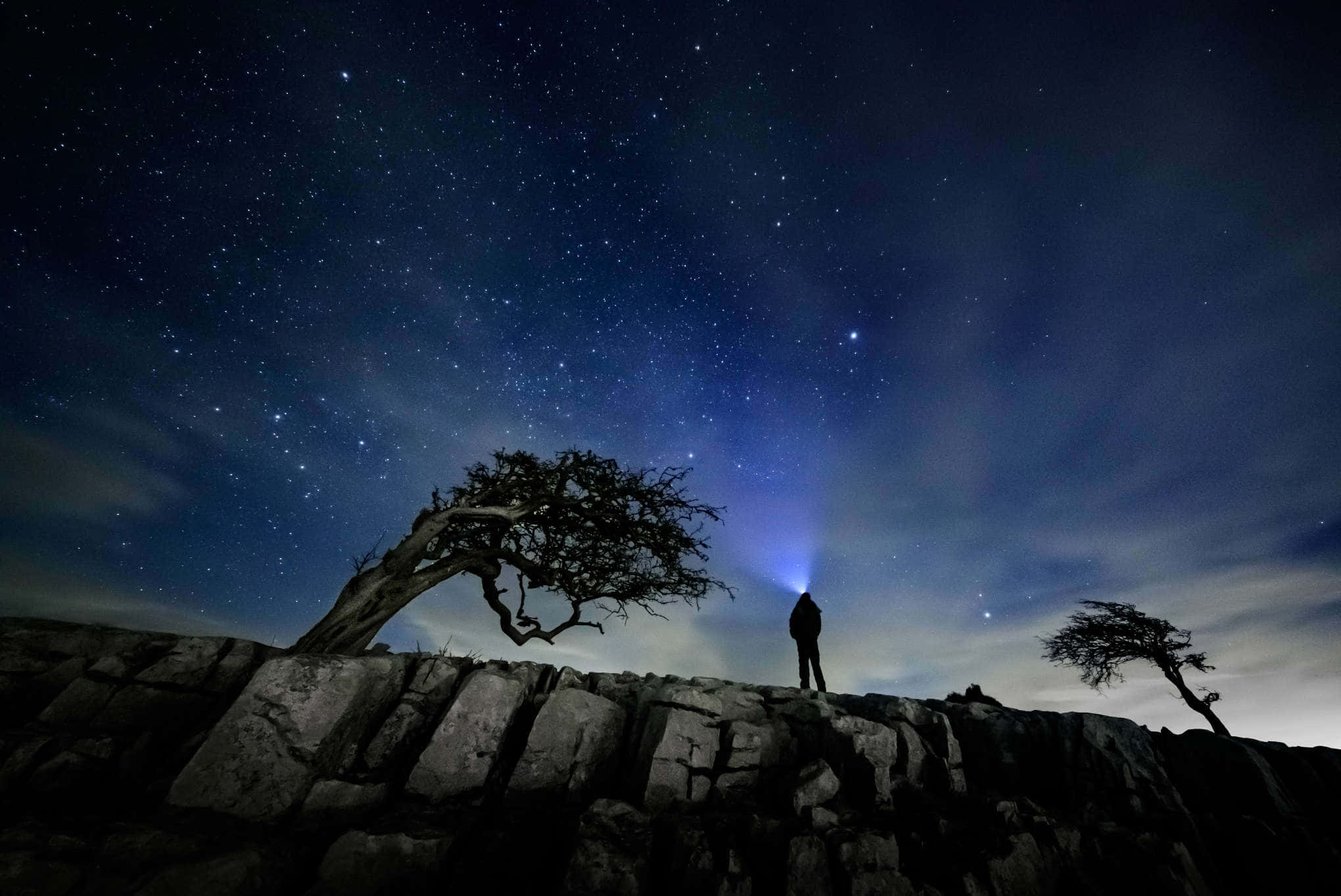 Stars 桌布 壁紙 — 夜晚，一個孤獨的身影站在岩石懸崖頂上，在星空背景下形成剪影，上方有明亮的藍色極光閃耀。